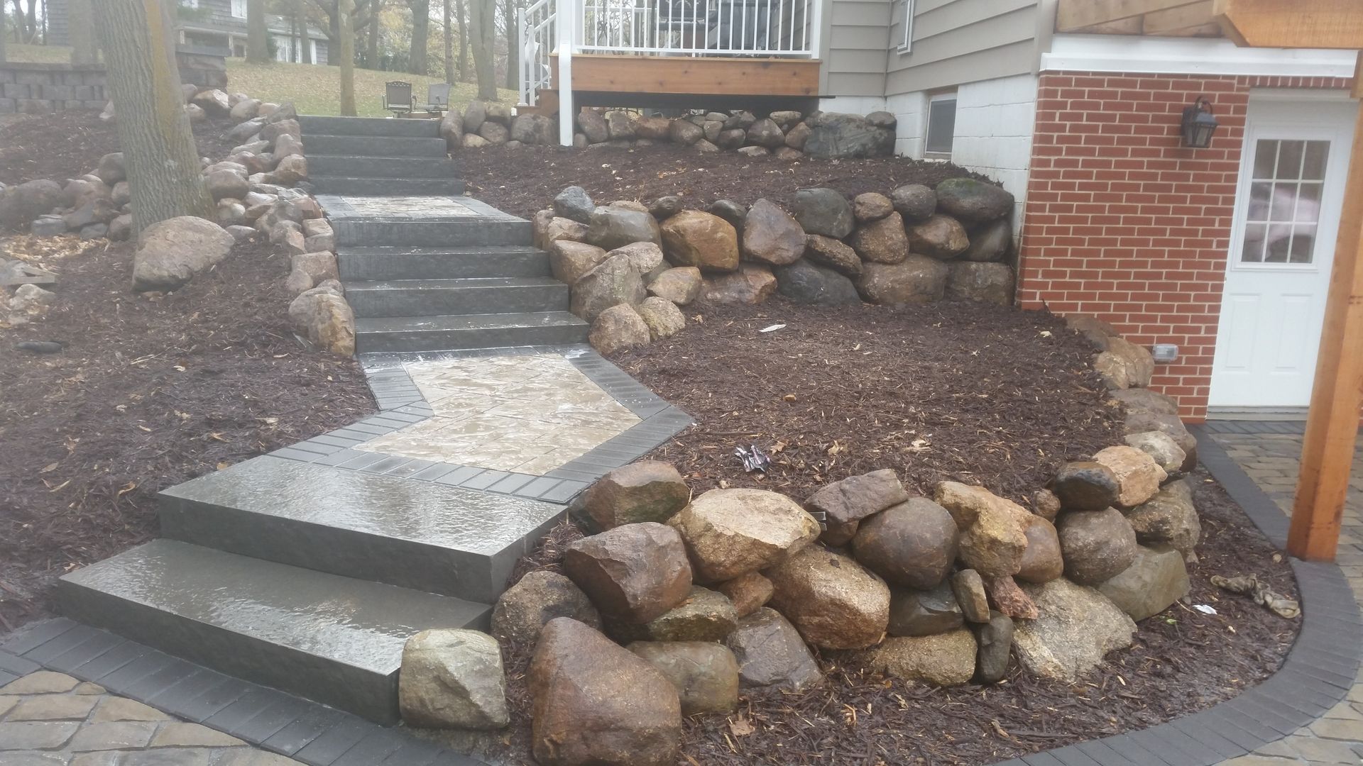 Stone stairs and a path wind through a garden with boulder retaining walls and mulch in front of a brick house.