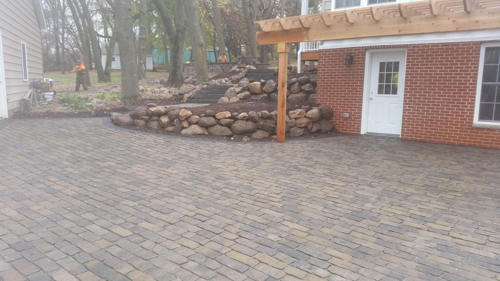 A patterned paver patio adjacent to a brick house with a white door and a stone retaining wall in a backyard.
