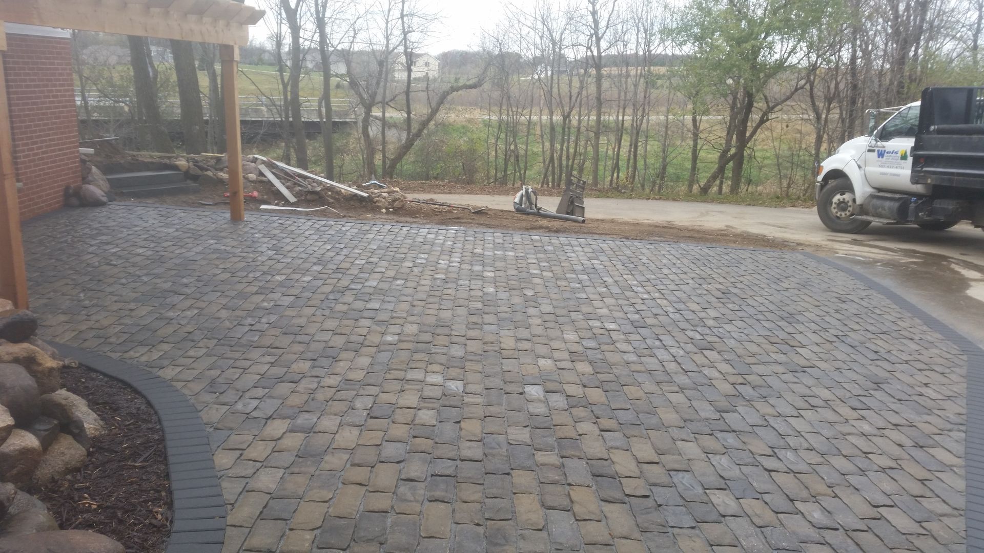 A freshly paved stone patio with a dark, arched border leading to a grassy yard and a parked utility truck.