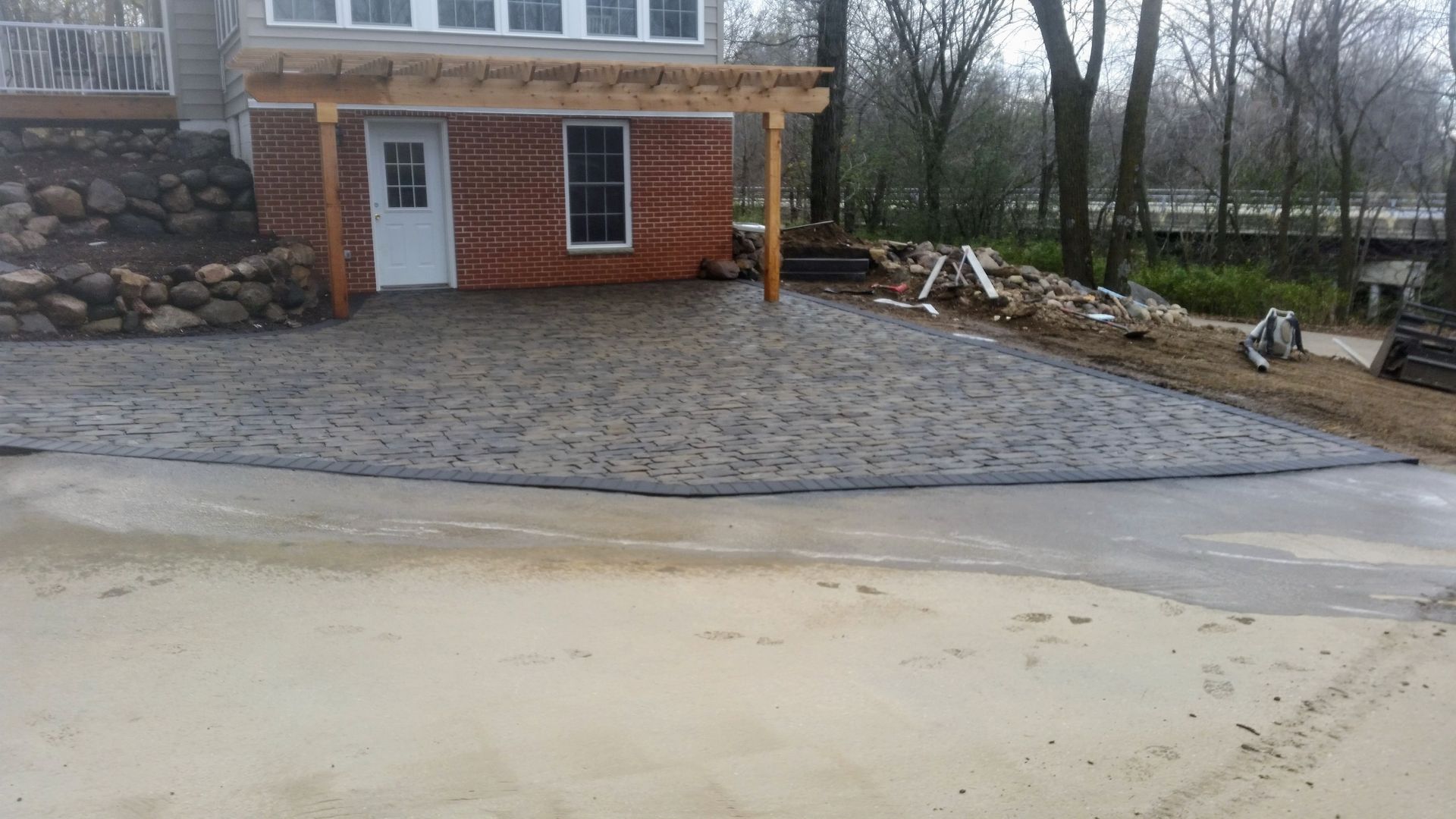 A newly installed grey stone patio adjacent to a brick house with a wooden pergola and an unfinished dirt yard.