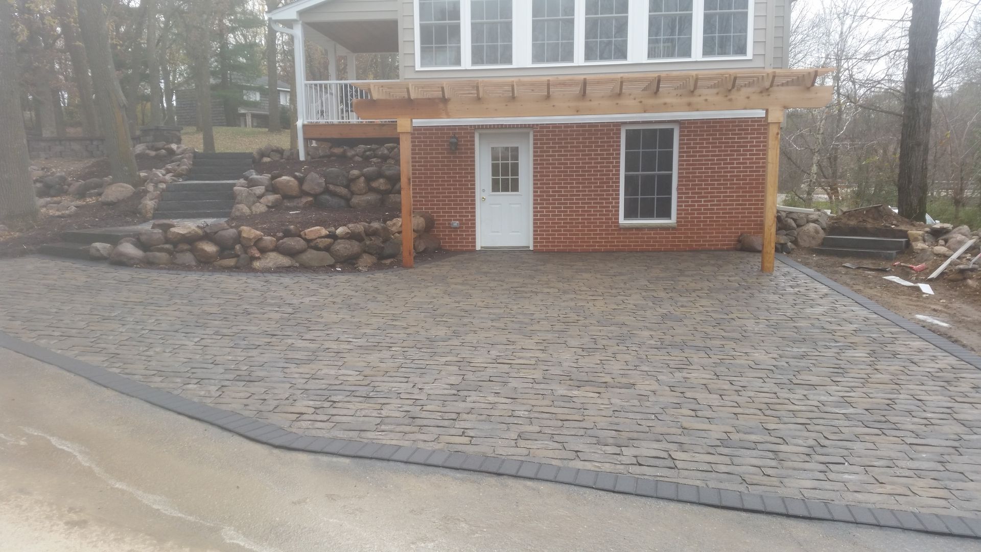 A paved patio with stone texture and a dark border, located in front of a brick house with a wooden pergola.