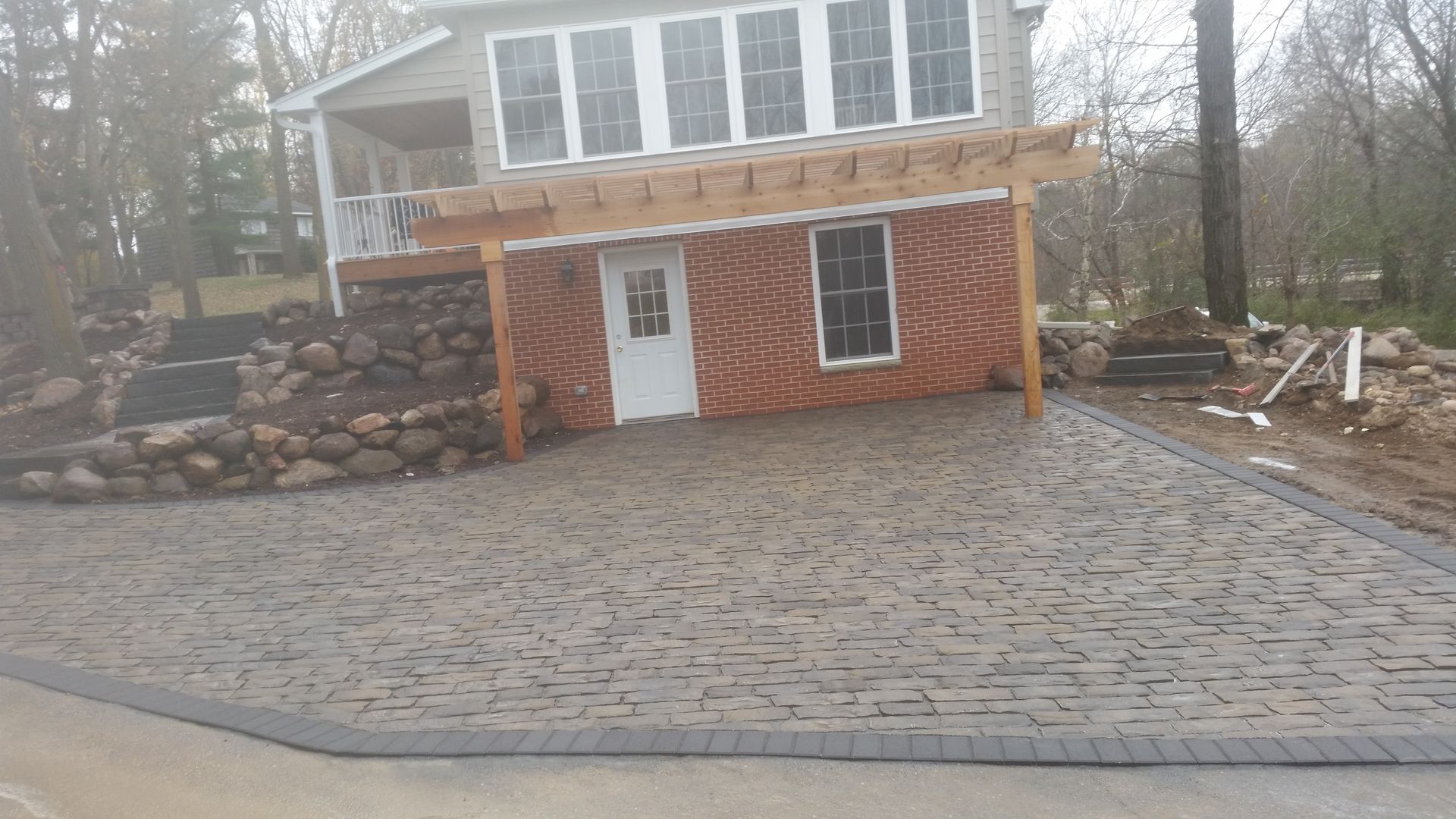 A multi-level backyard with a patterned stone patio, a wooden pergola, a tiered rock wall, and a house under renovation.