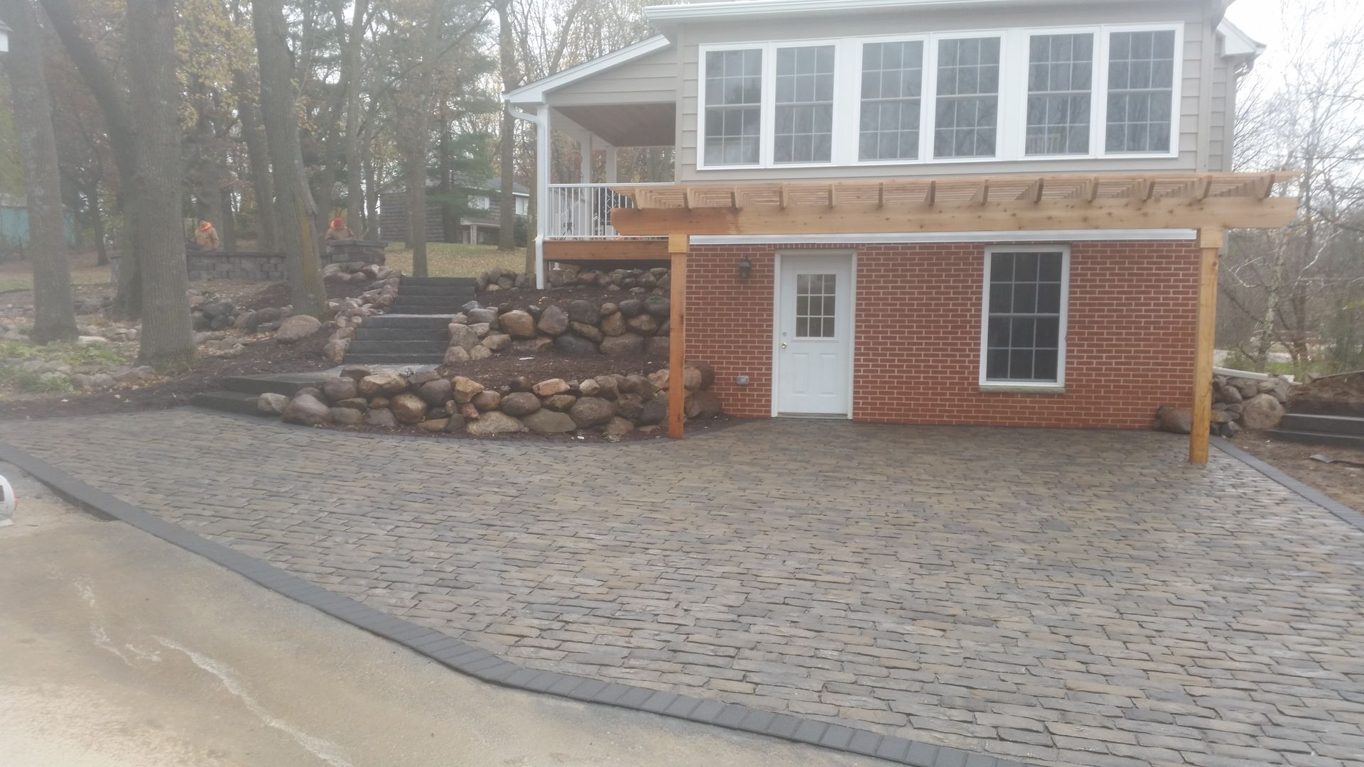 A house with a large stone-paver patio, a rustic stone retaining wall, and a wooden pergola over a brick-faced lower level.