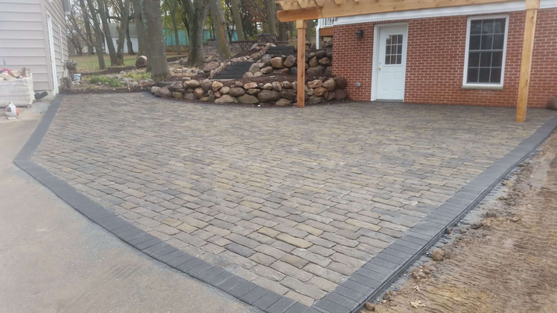 A newly installed gray paver patio with a dark charcoal border, situated beside a brick building and a stone retaining wall.