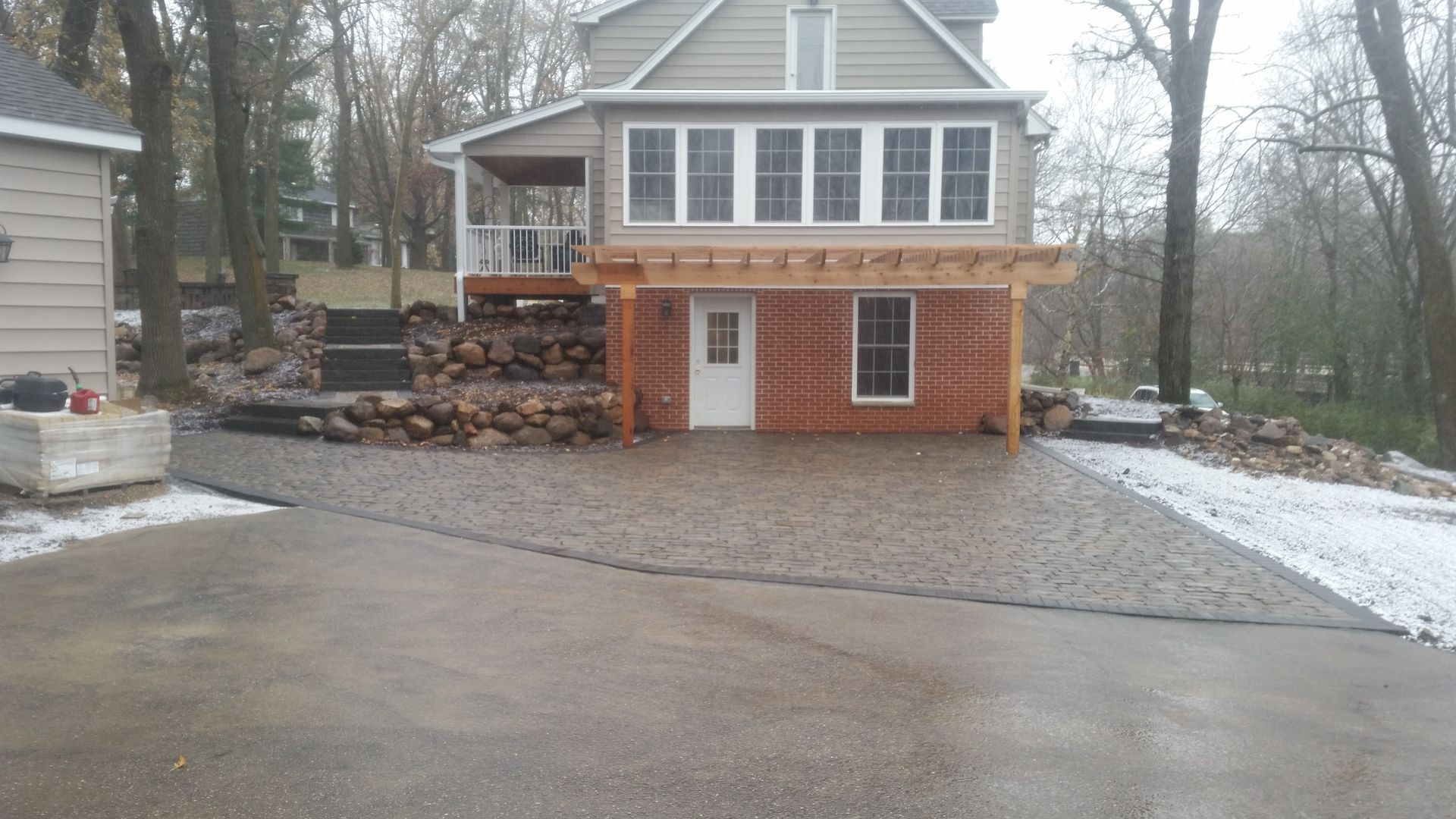 A two-story house with a brick ground floor, paved patio, and wooden pergola, surrounded by trees on a cloudy, winter day.