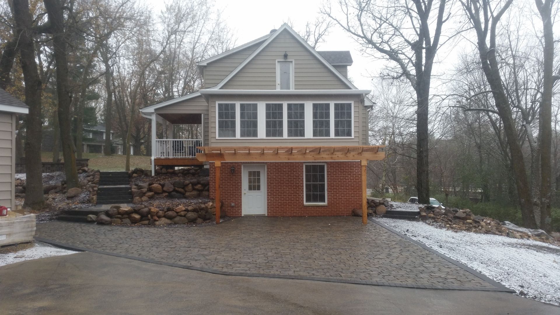 A two-story house with a brick ground floor, grey upper level, large deck, and a stone-paved driveway in a wooded area.