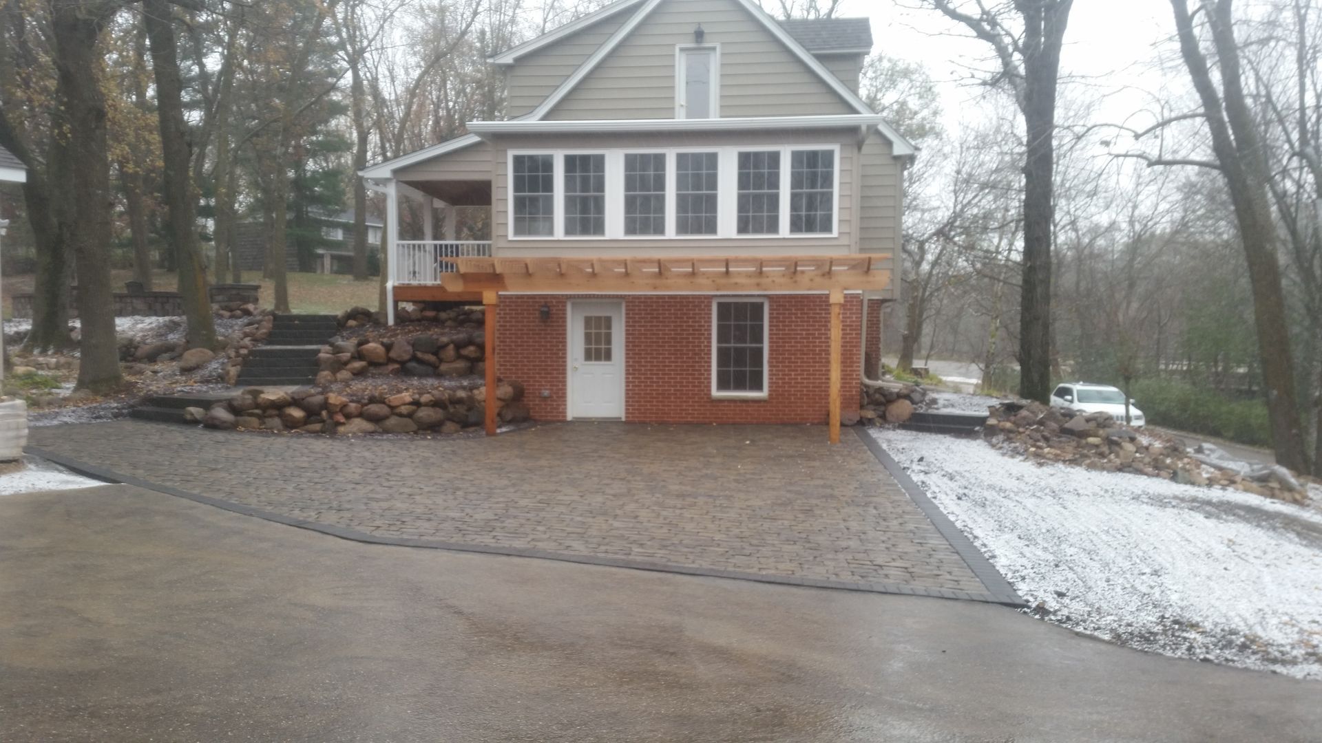 A two-story house with a white door, a red brick base, and a new gravel driveway in a wooded, partially snowy setting.