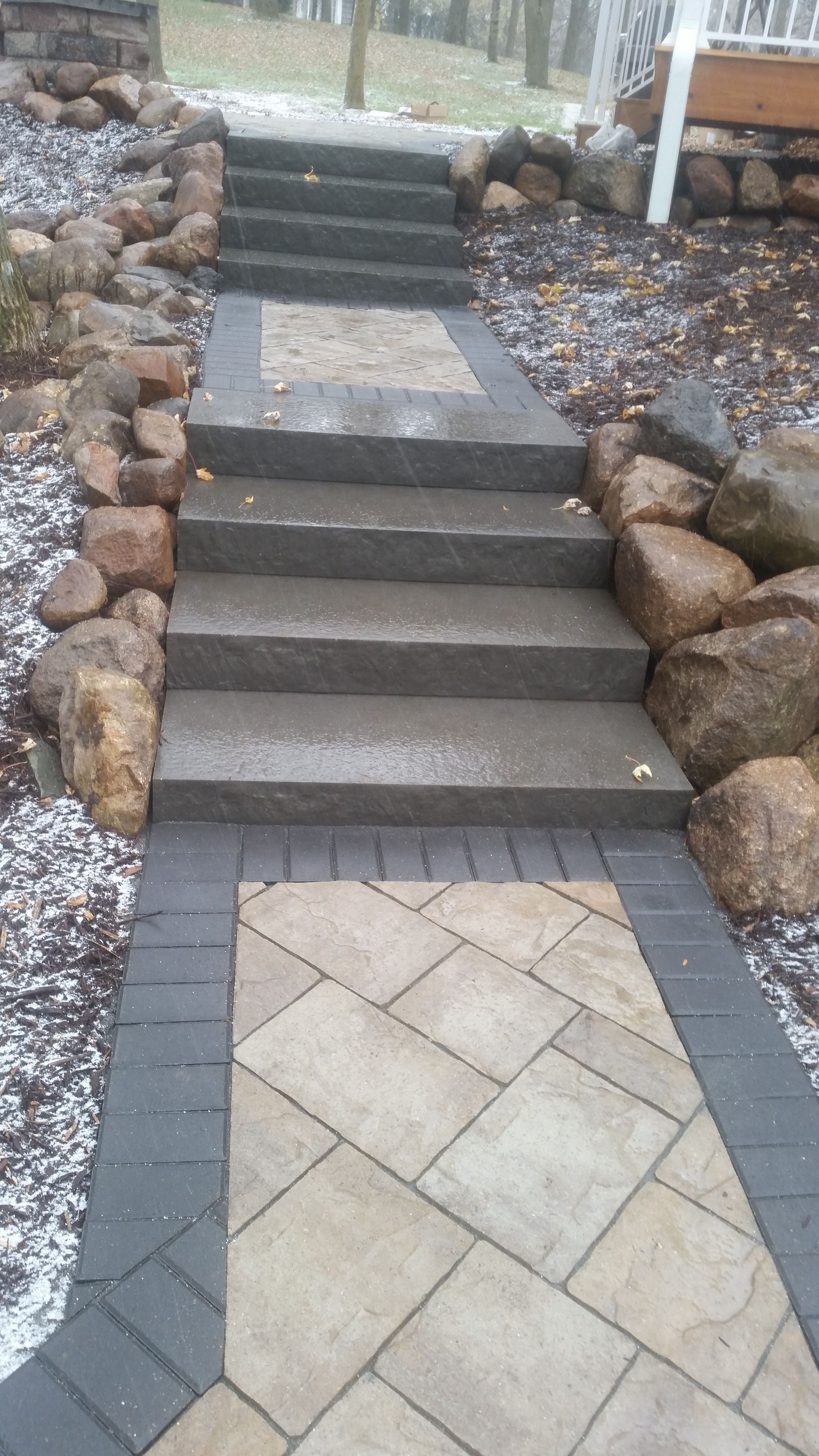 Outdoor staircase made of dark stone steps, bordered by large boulders and a paved walkway with a tan and dark gray pattern.