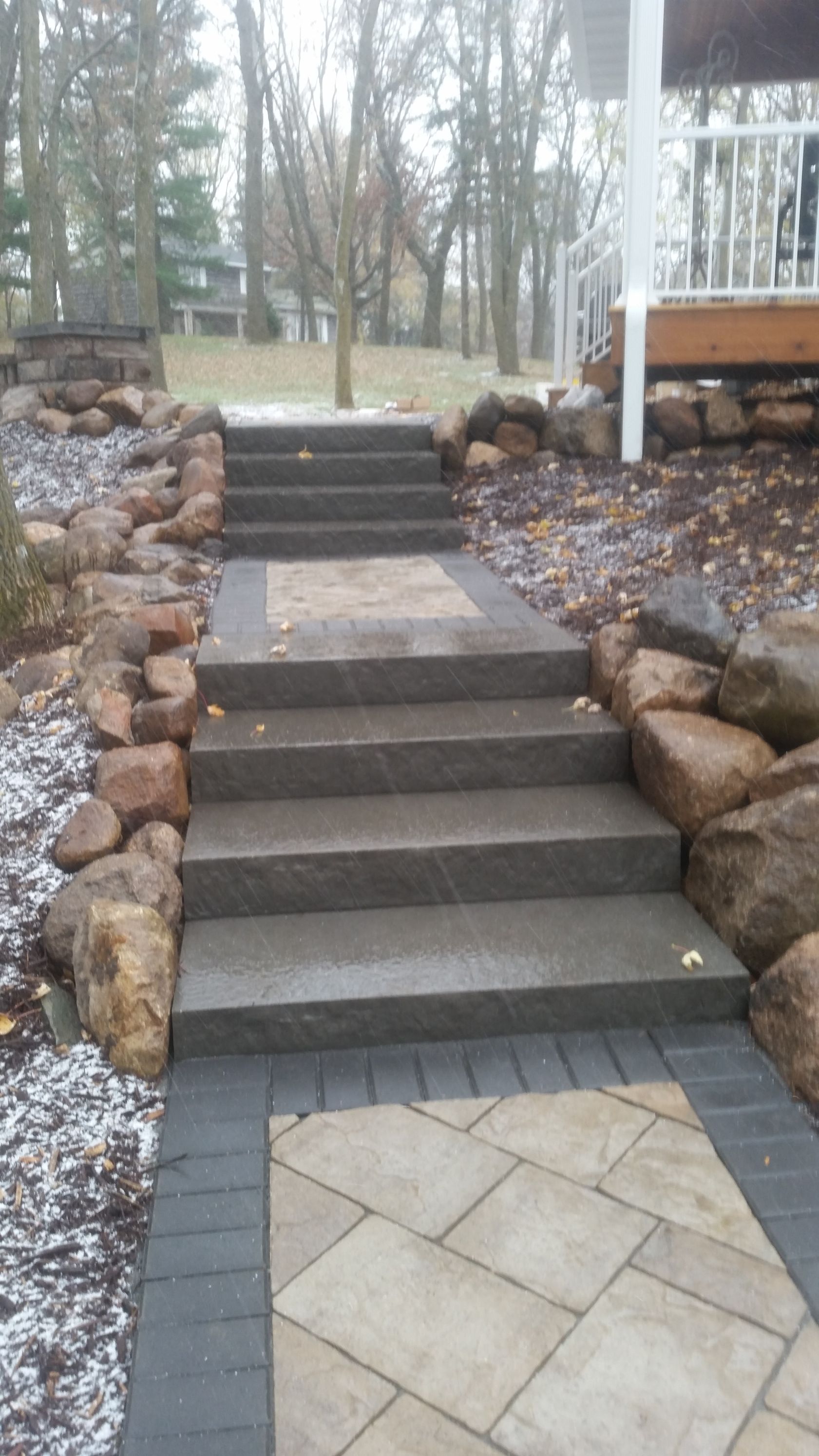 A stone staircase leads up to a deck, flanked by rock retaining walls and a paved walkway in a wooded yard.