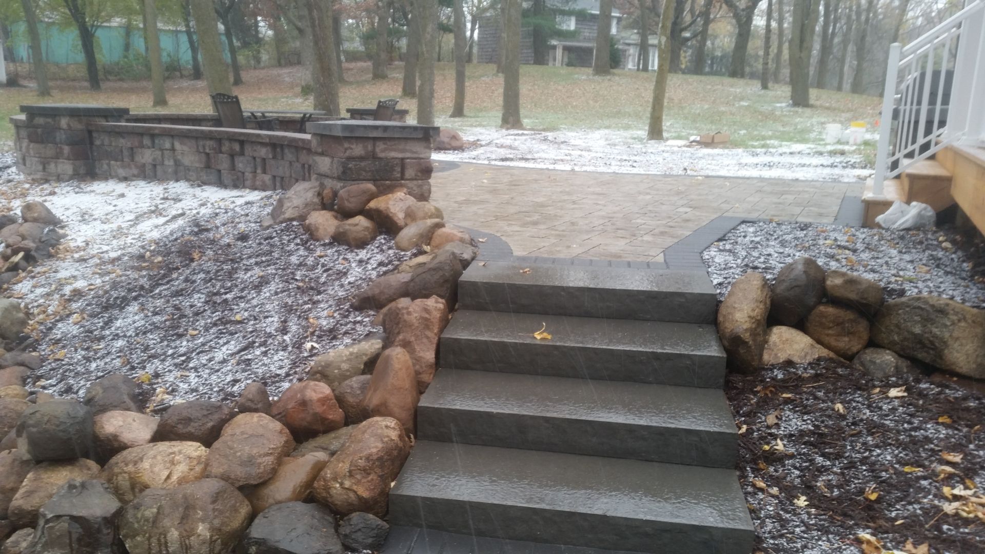 Concrete steps lead to a stone patio with a low wall, surrounded by boulders and light snow, beside a white porch railing.