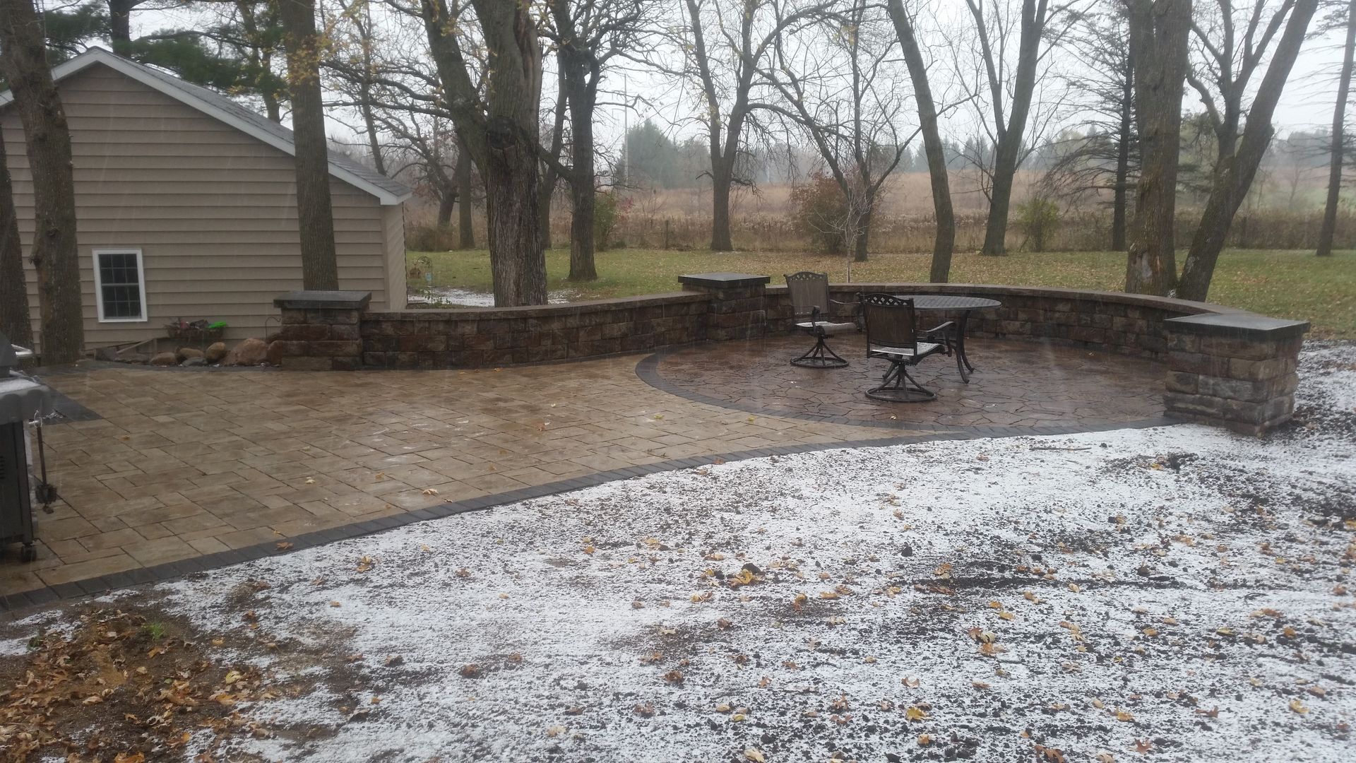 A stone patio with a curved retaining wall and outdoor furniture on a lightly snow-covered lawn beside a house.