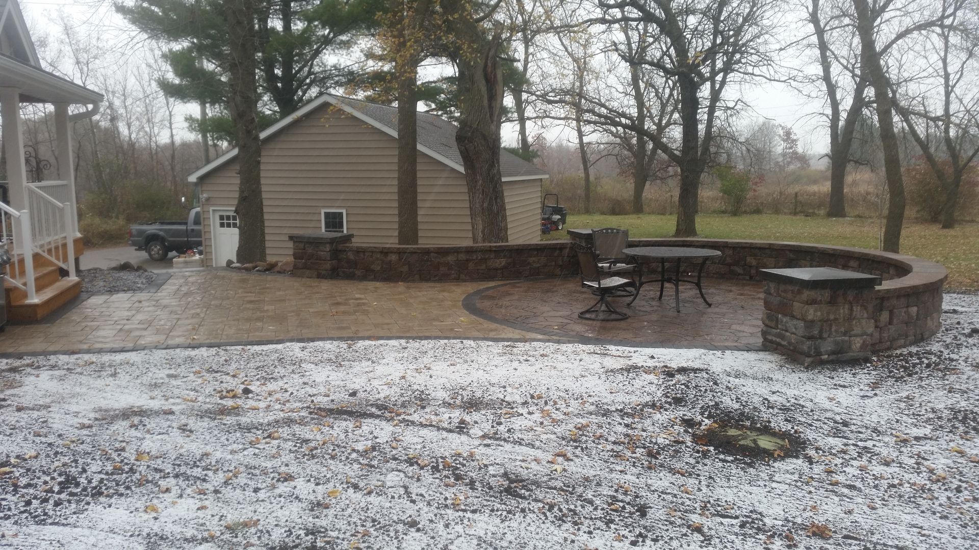 A stone patio with a curved retaining wall, a small table, and chairs in a yard dusted with light snow.