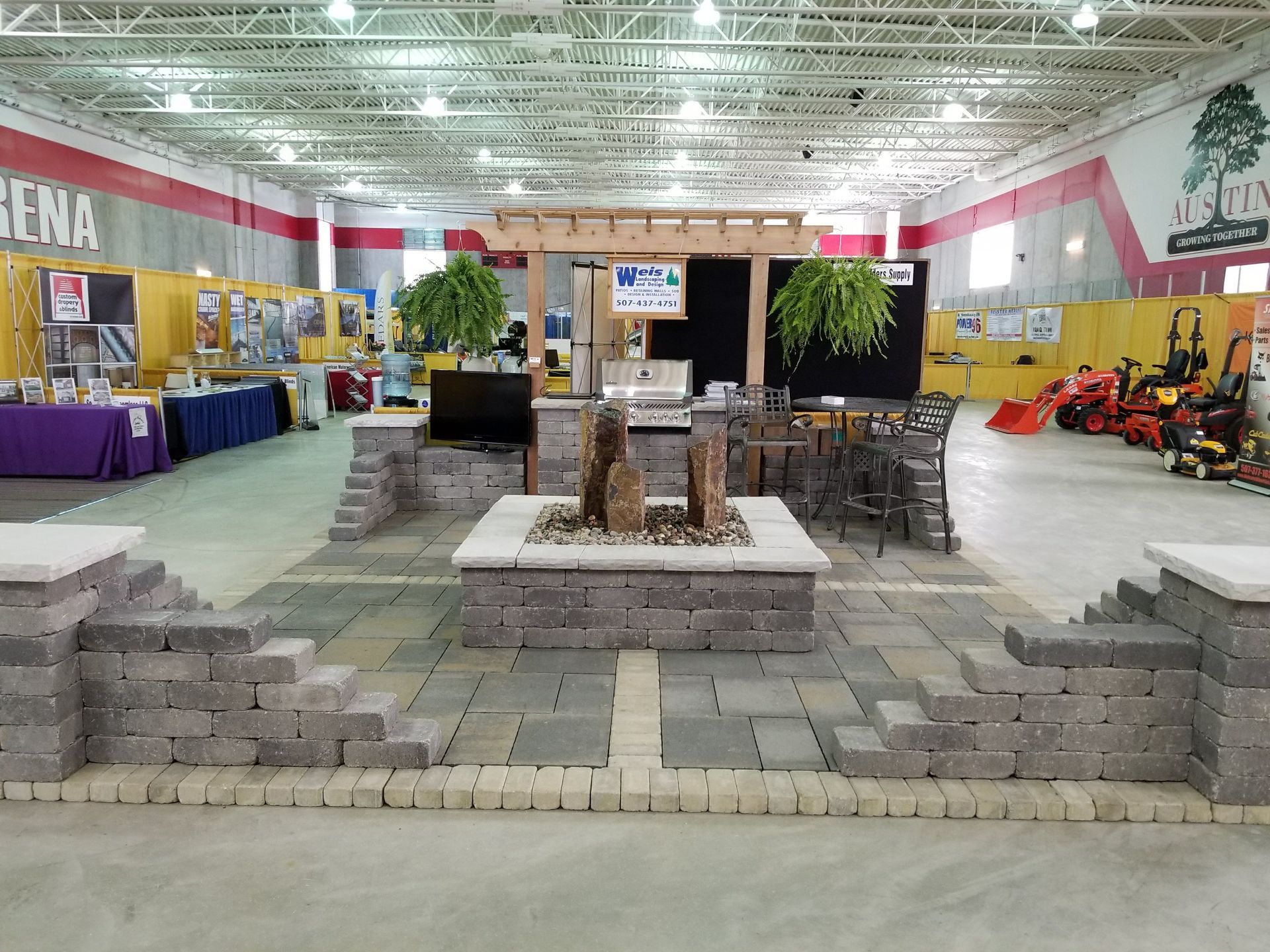 A display of gray stone patio pavers, a fire pit, and a wooden pergola set up inside a large trade show arena.