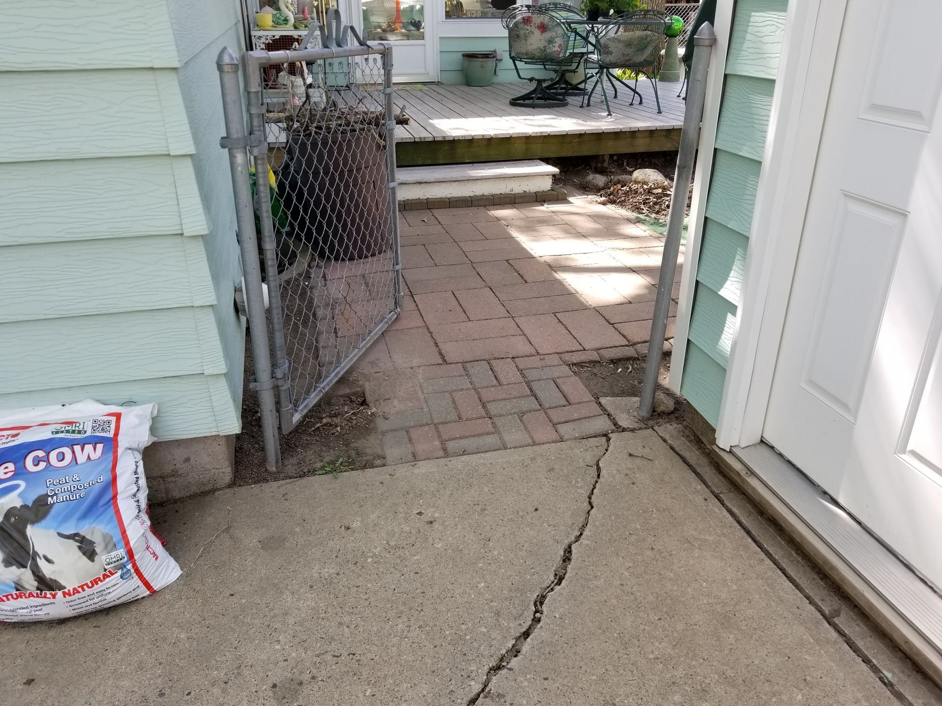 A cracked concrete walkway leading to a brick path and an open chain-link gate between two houses.