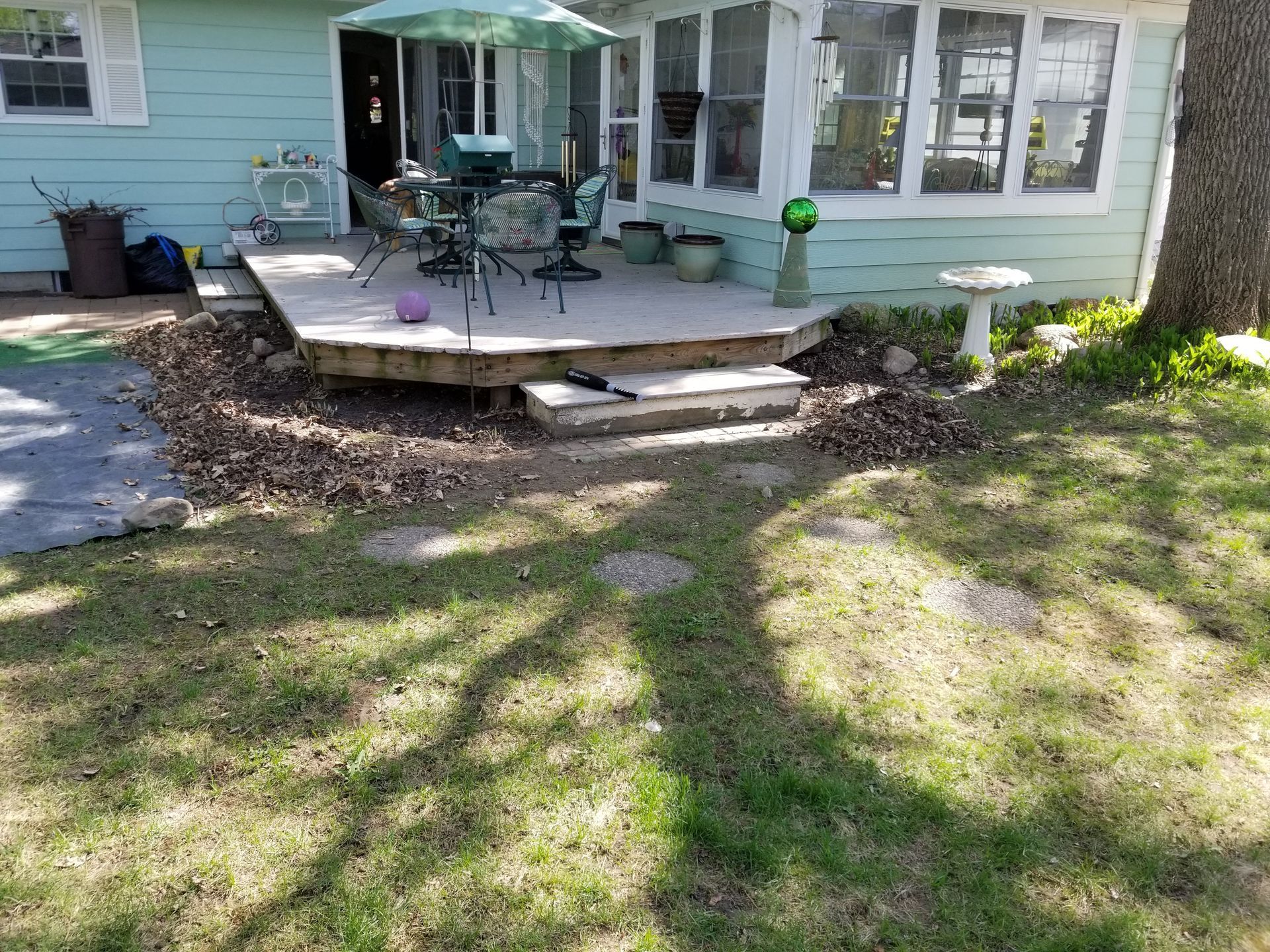 Light blue house with a wooden patio, outdoor table and chairs under a green umbrella, and a birdbath in a grassy yard.