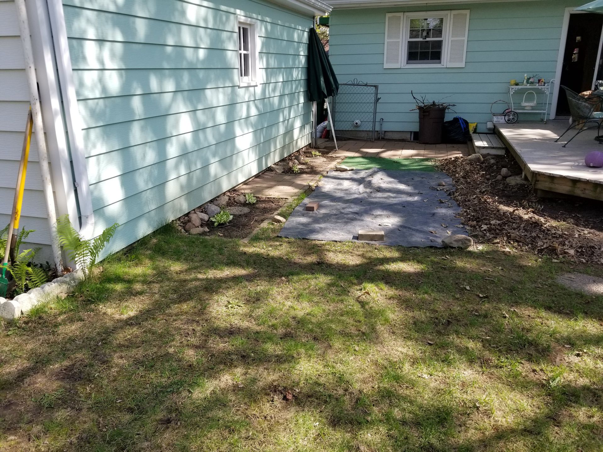A side yard with light blue siding, a patch of bare ground covered by a tarp, and a wooden deck.