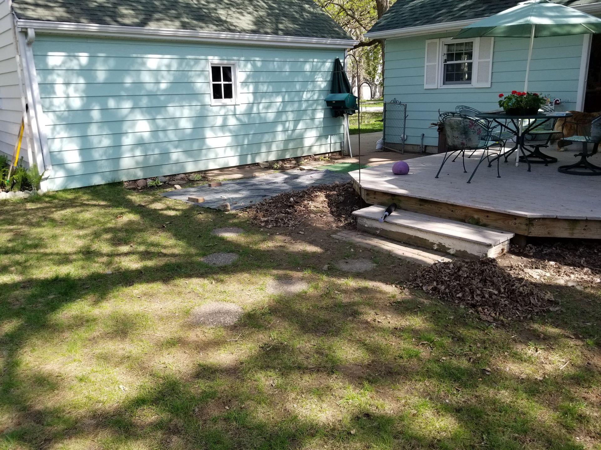 A light blue garage and house with a wooden deck, a patio set, a green umbrella, and a grassy yard with stone pavers.