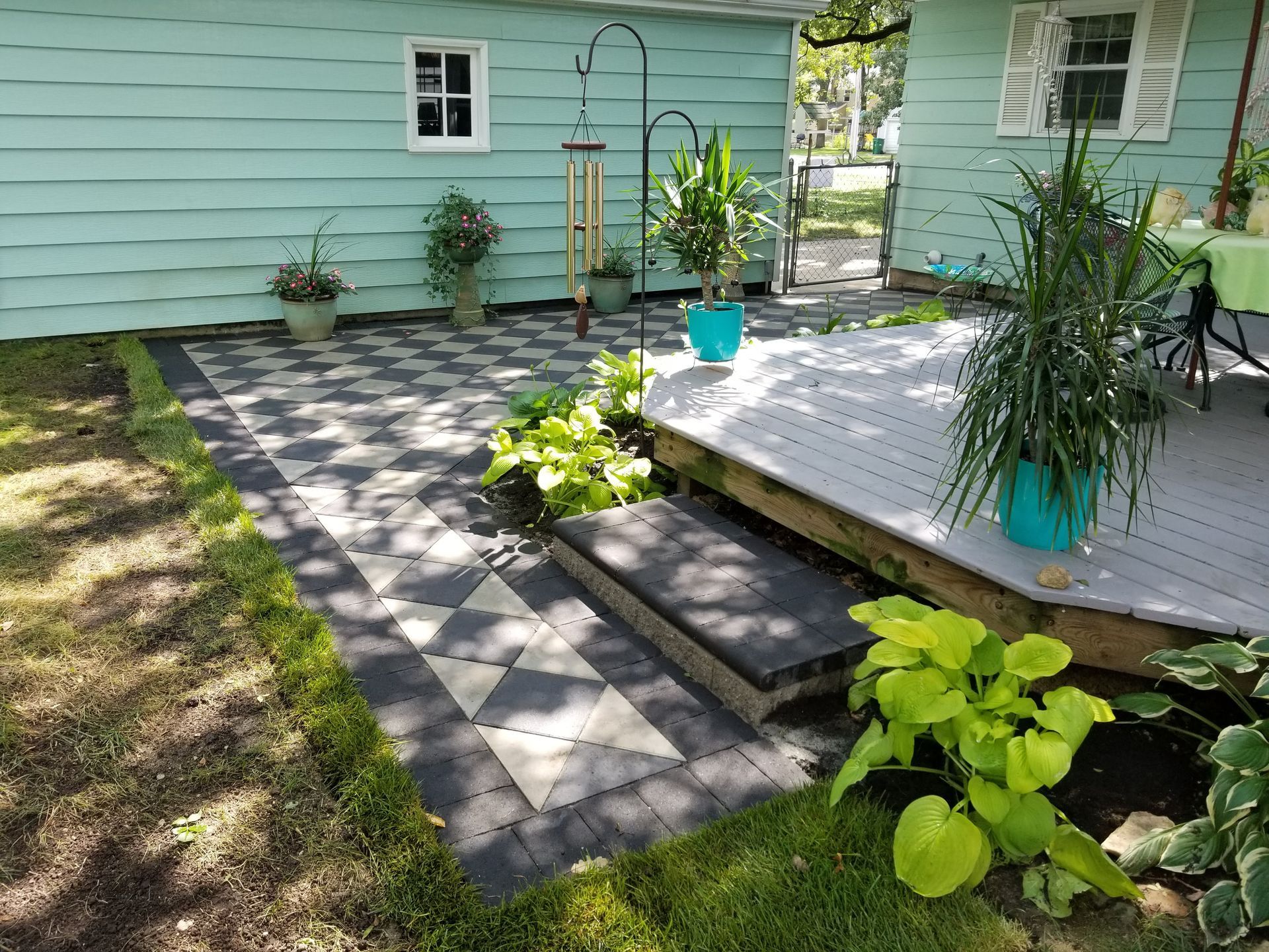 A light blue house with a geometric gray and black paver patio, a wooden deck, and several potted plants.