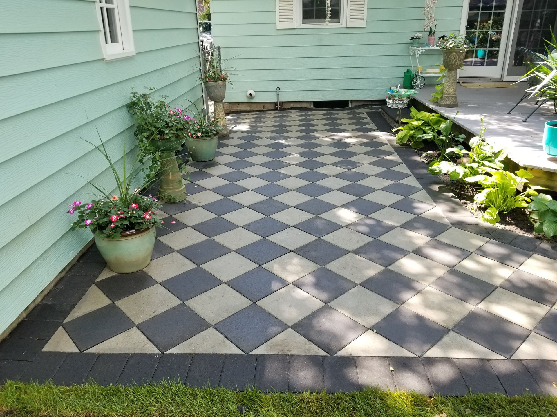 A patio with a gray and black checkerboard paver design, bordered by potted plants next to a light green house.