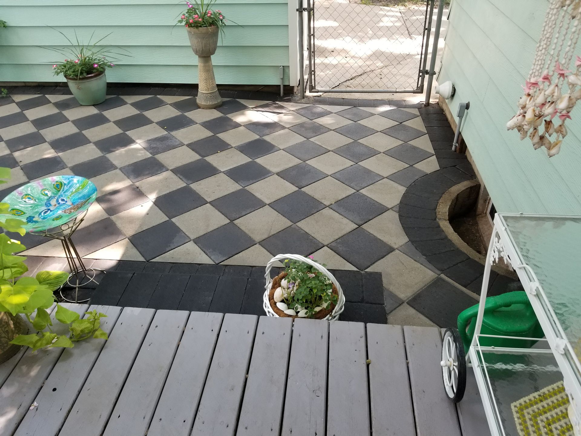A patio with a gray wooden deck in the foreground and a black-and-white checkered paver area beyond it, featuring planters.