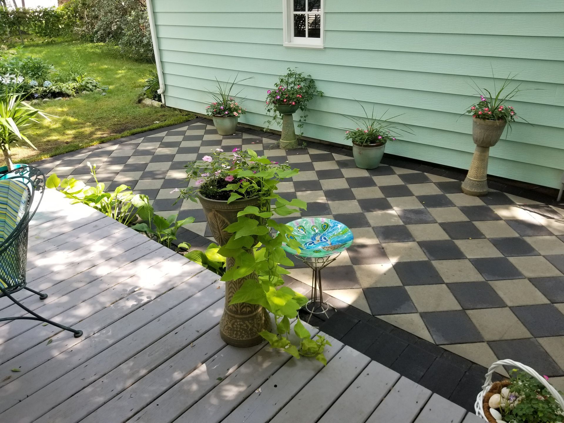 A green house with a gray deck leading to a patio with black and gray checkered pavers, potted plants, and a birdbath.