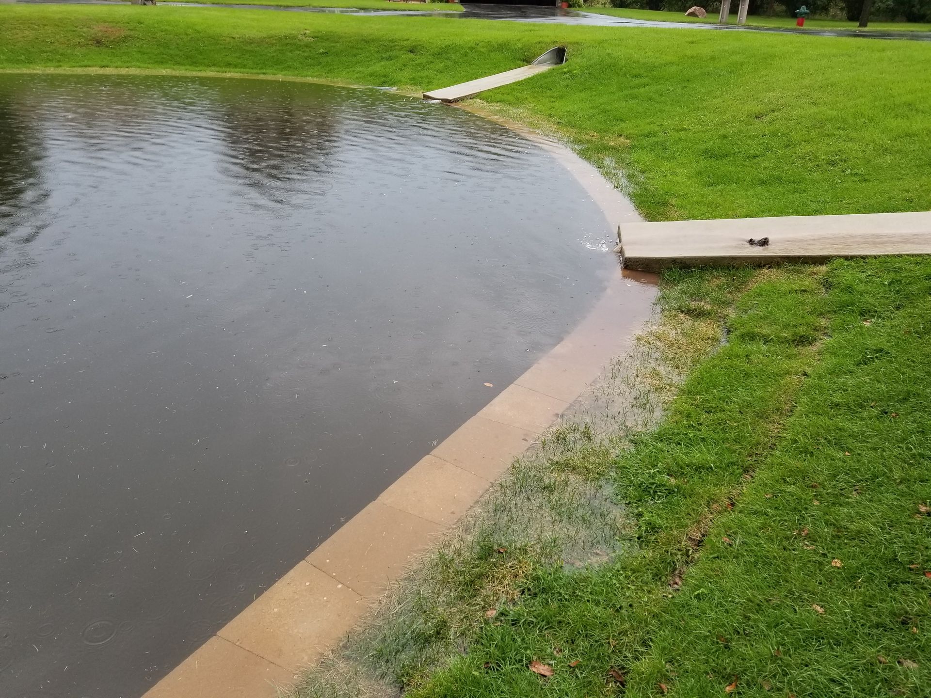 A pond edge featuring a concrete border and two concrete drainage pipes discharging water into the grass and pond.