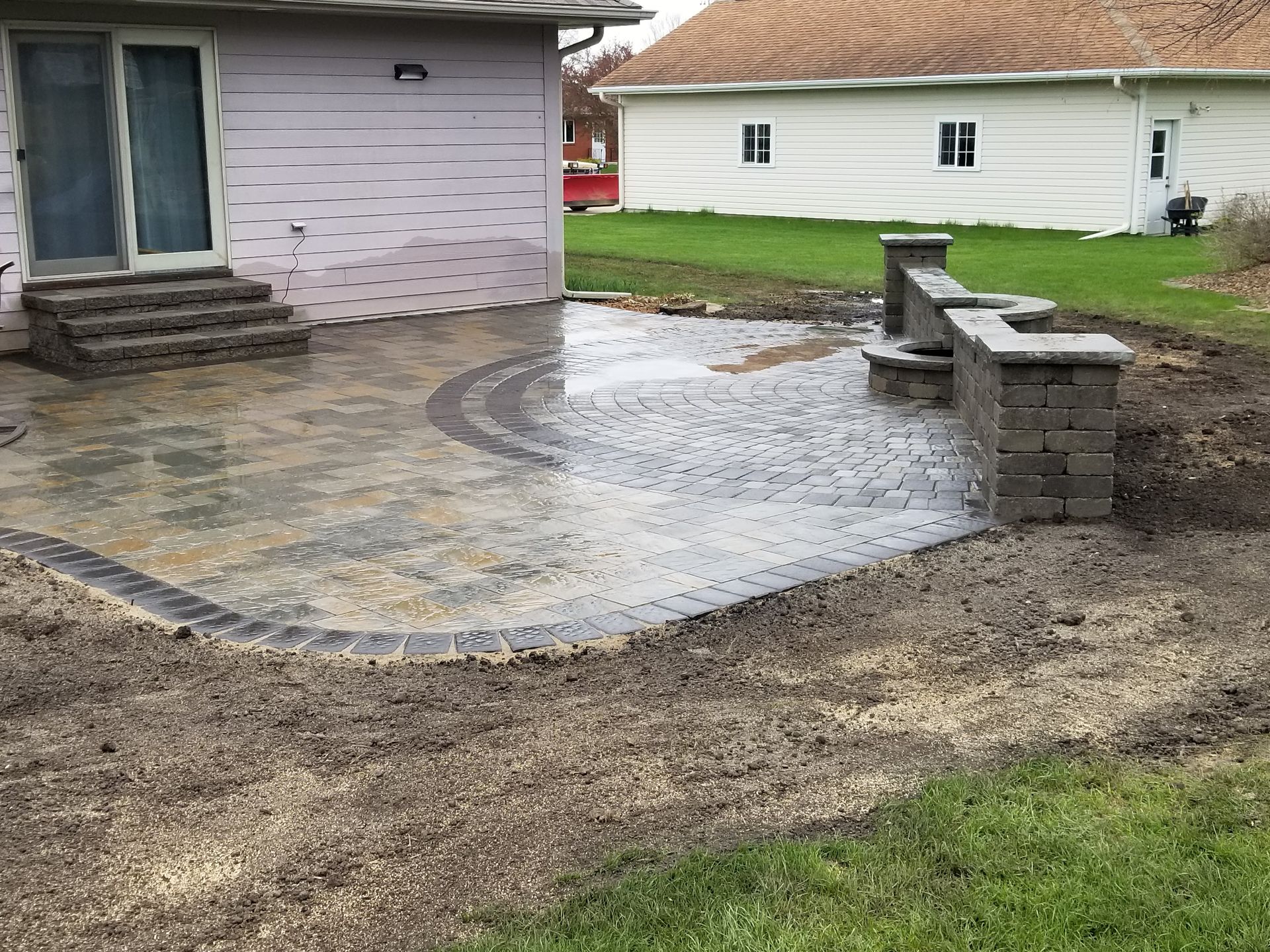 A new stone patio with a curved wall and steps leading to a house, surrounded by unfinished dirt and lawn.