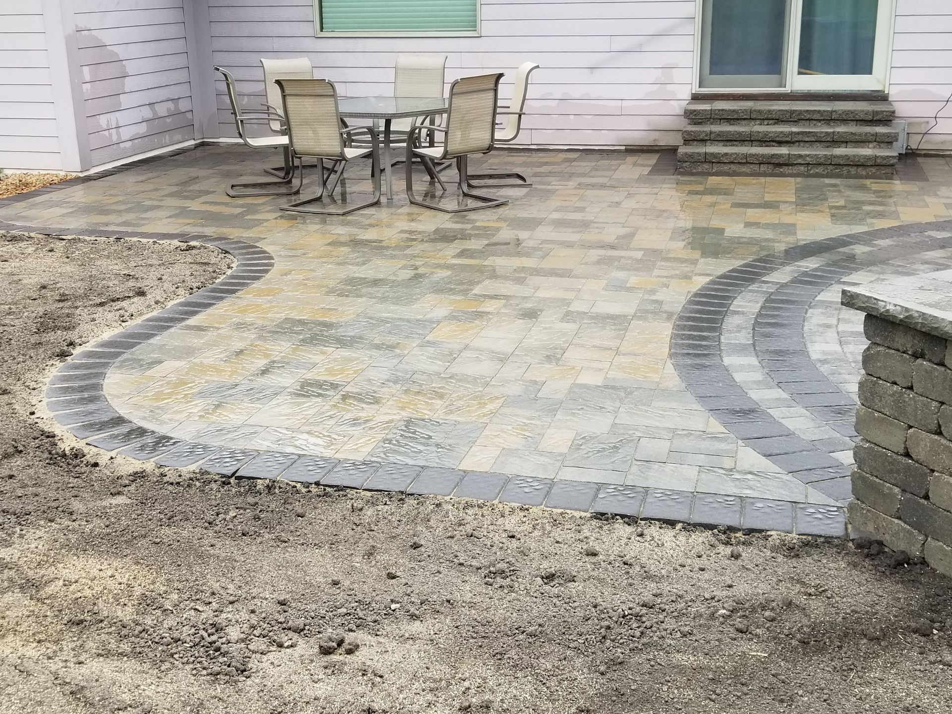 A newly installed stone paver patio with a curved dark border, set against a house exterior with patio furniture.