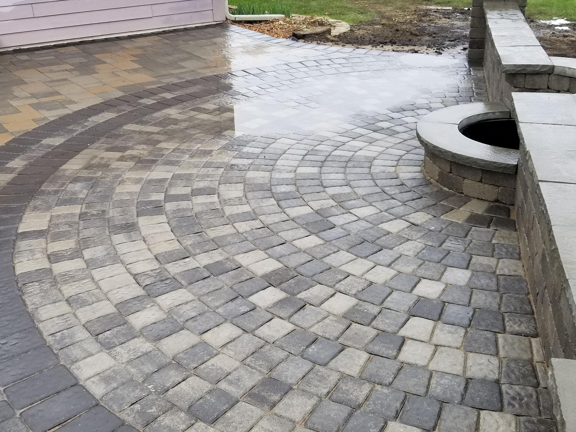 A patio made of grey and tan stone pavers arranged in a circular pattern, with a built-in stone fire pit and wall.