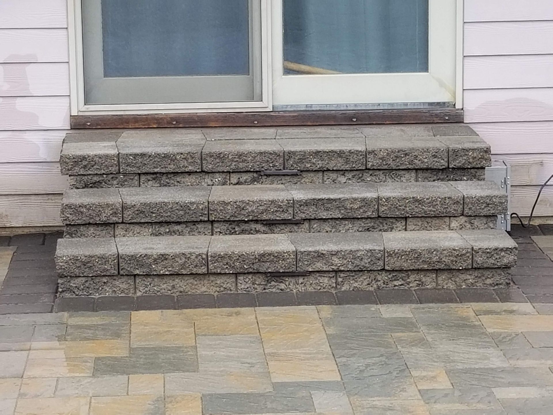 Three stone steps leading up to a sliding glass door against a light-colored house exterior with a paved patio.