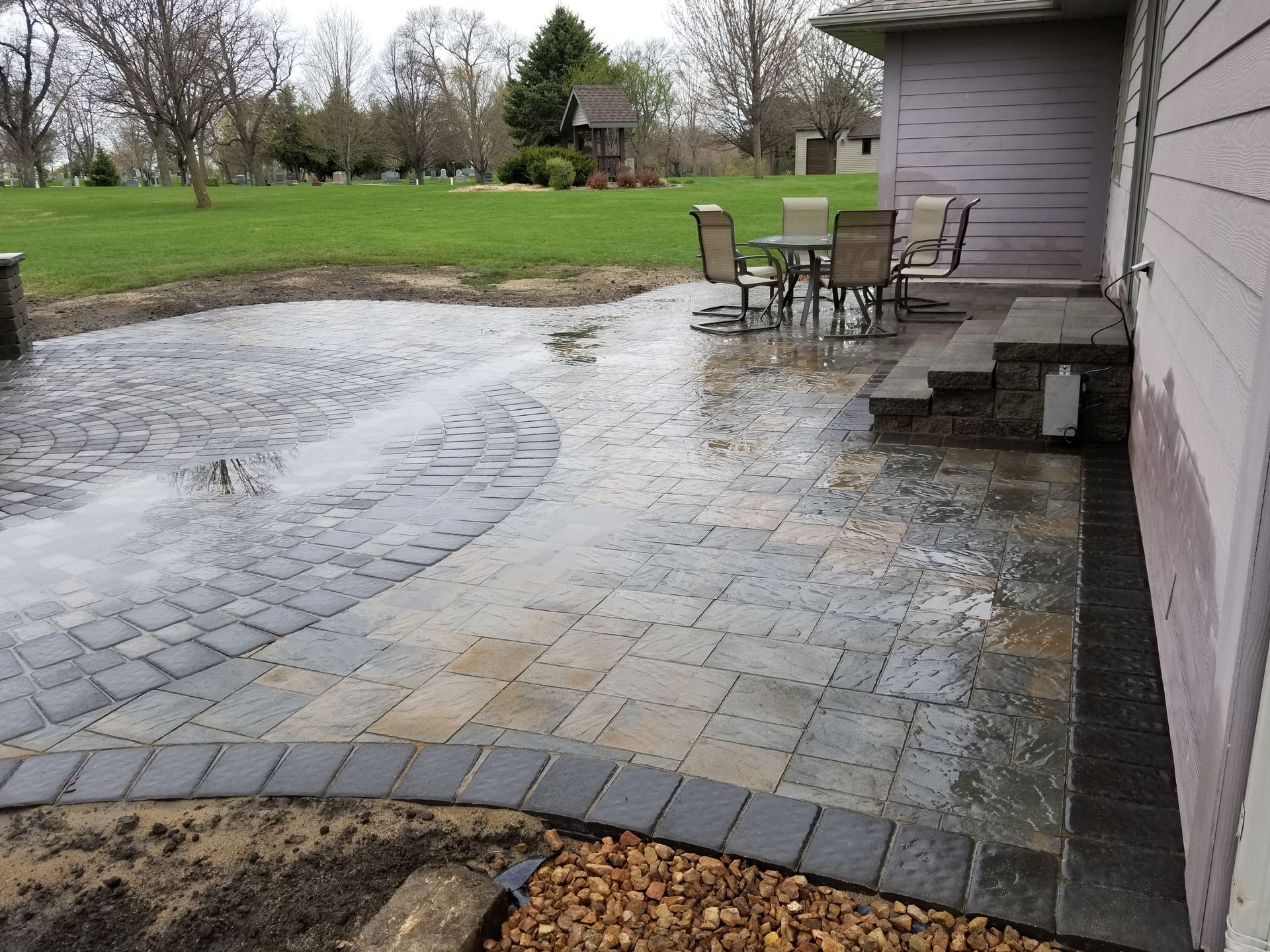 A patio with gray and brown interlocking stone pavers, set with a table and four chairs next to a house exterior.