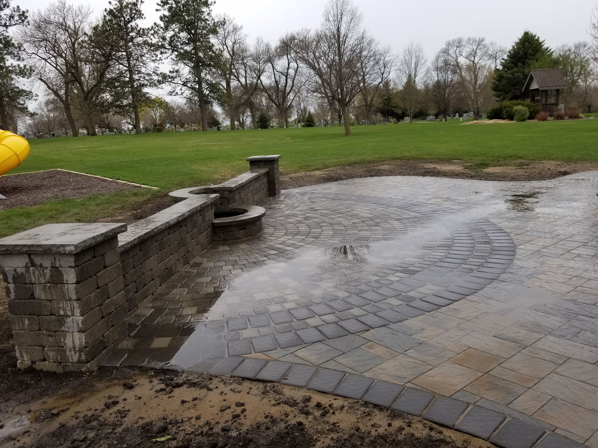 A newly paved stone patio with a curved retaining wall and fire pit, situated in a backyard with a large grass lawn.