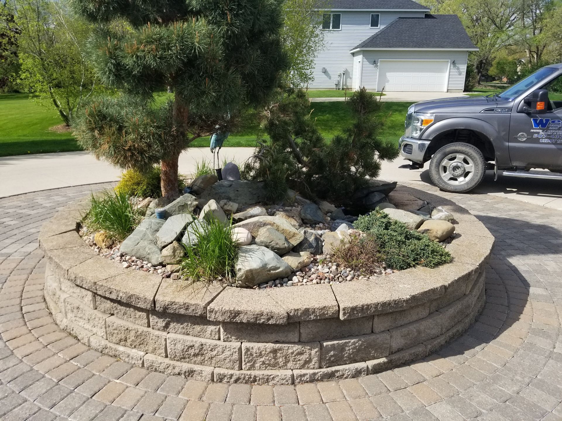 A circular stone planter with a small pine tree and rock landscaping sits in the center of a paved driveway.