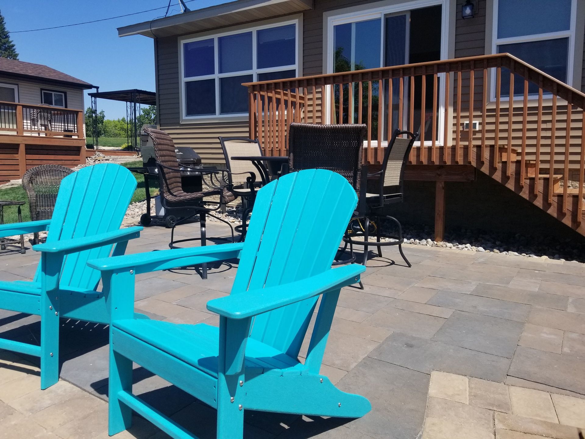 Two bright turquoise Adirondack chairs on a stone patio in front of a house with a wooden deck.