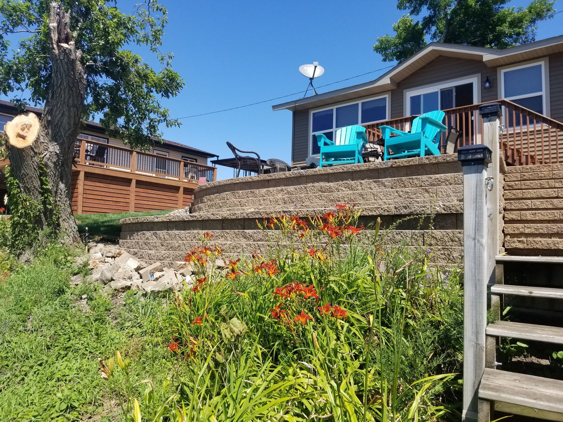 A landscaped garden with bright orange flowers in front of a tiered retaining wall, a patio with chairs, and a house.