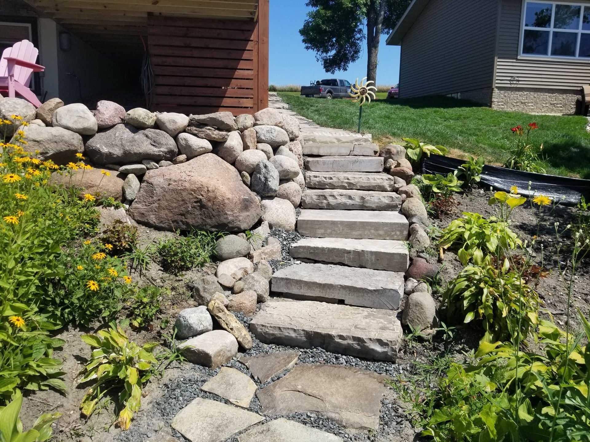 Stone steps leading up a grassy embankment next to a brown wood-paneled building and a house with green siding.