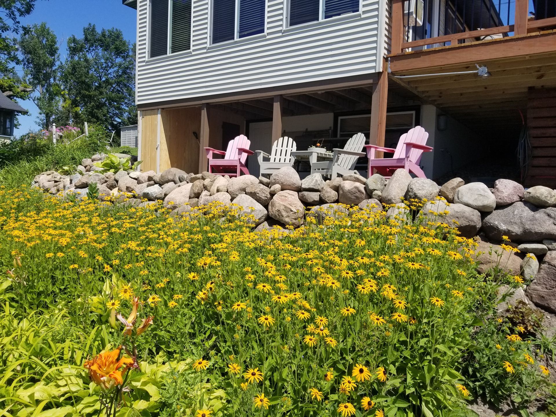 A house with a stone retaining wall and a field of yellow flowers, featuring a deck with several outdoor chairs underneath.