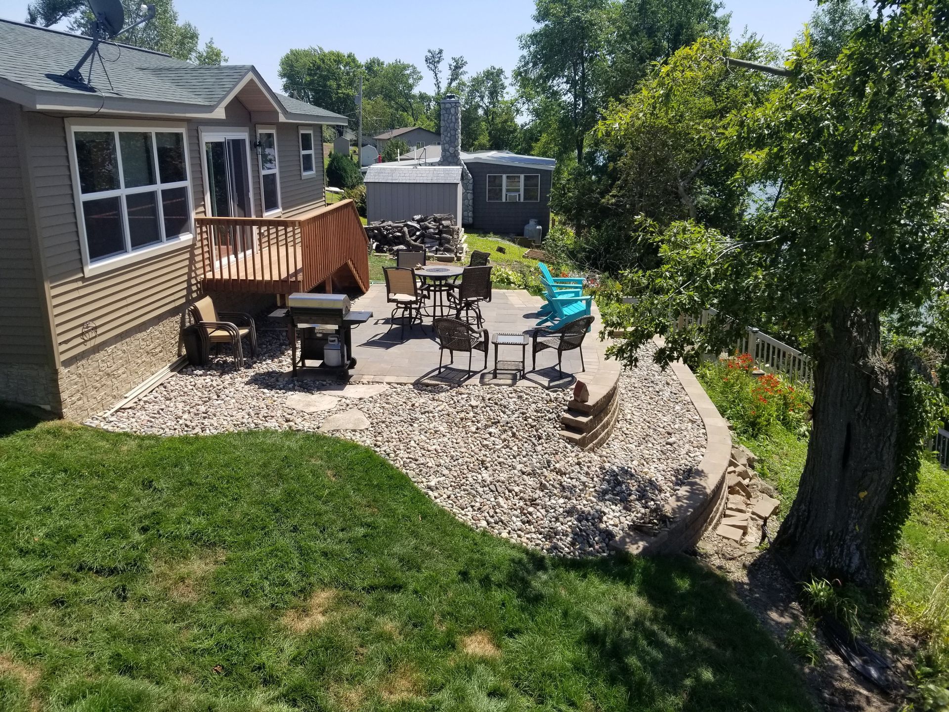 A house with a raised wooden deck and a stone patio area with outdoor furniture and a grill overlooking a lake.