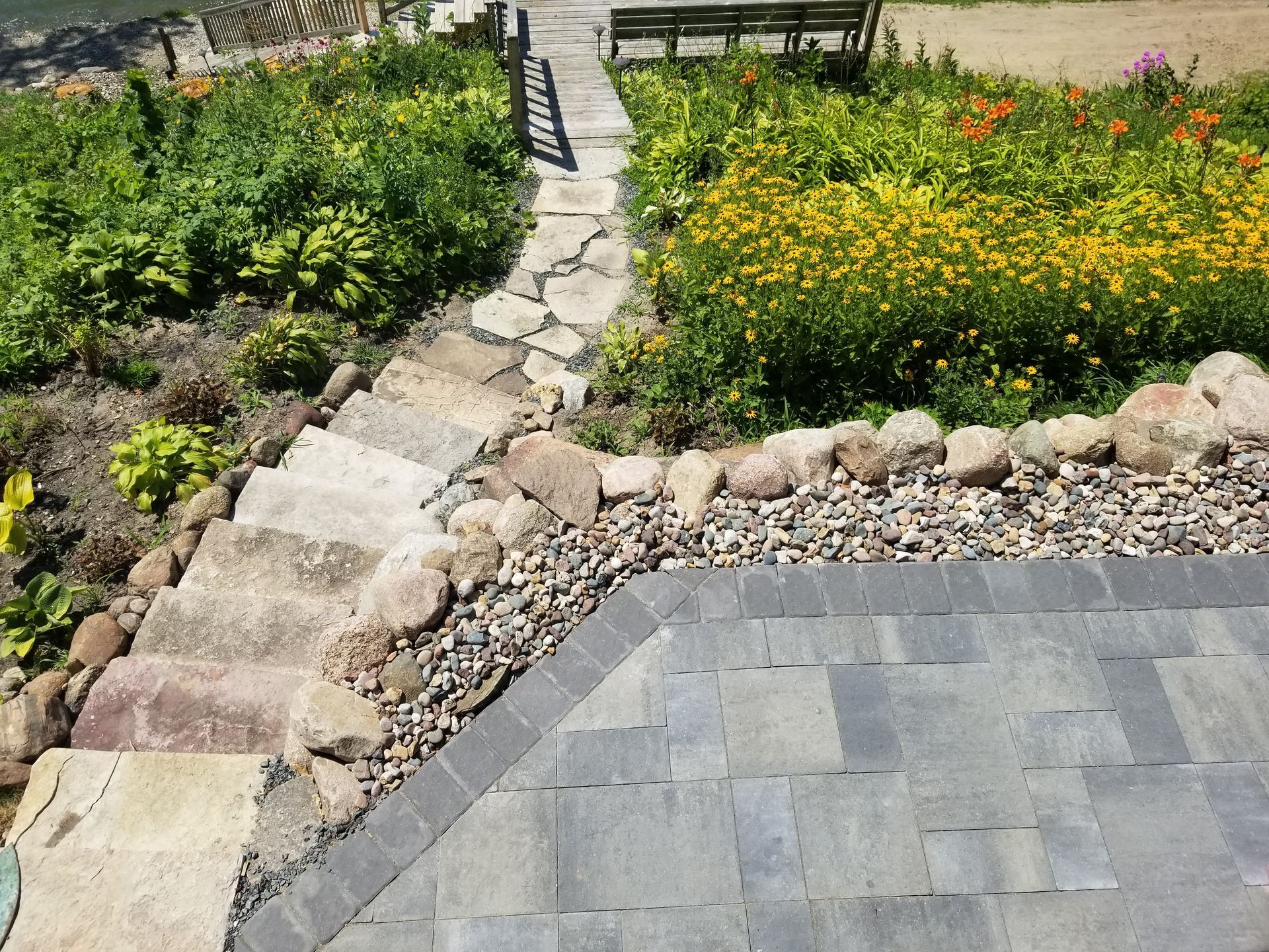 A stone stairway leading from a gray paver patio up into a sunny garden with yellow flowers and a wooden bench.