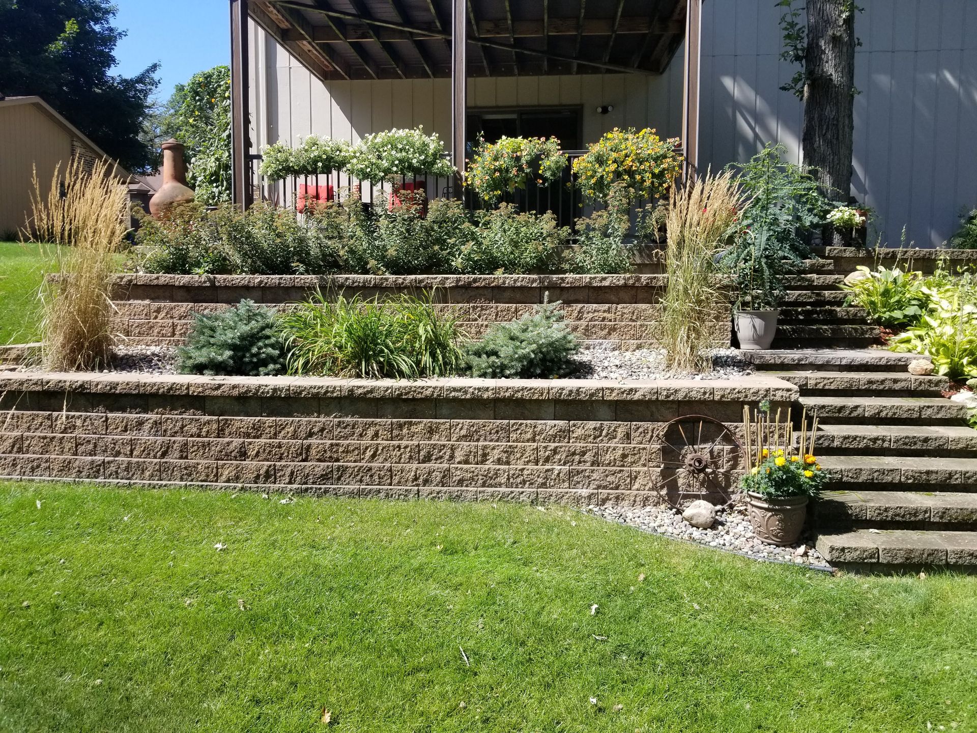 Tiered stone retaining walls with garden beds, ornamental grasses, and concrete steps leading to a backyard deck.
