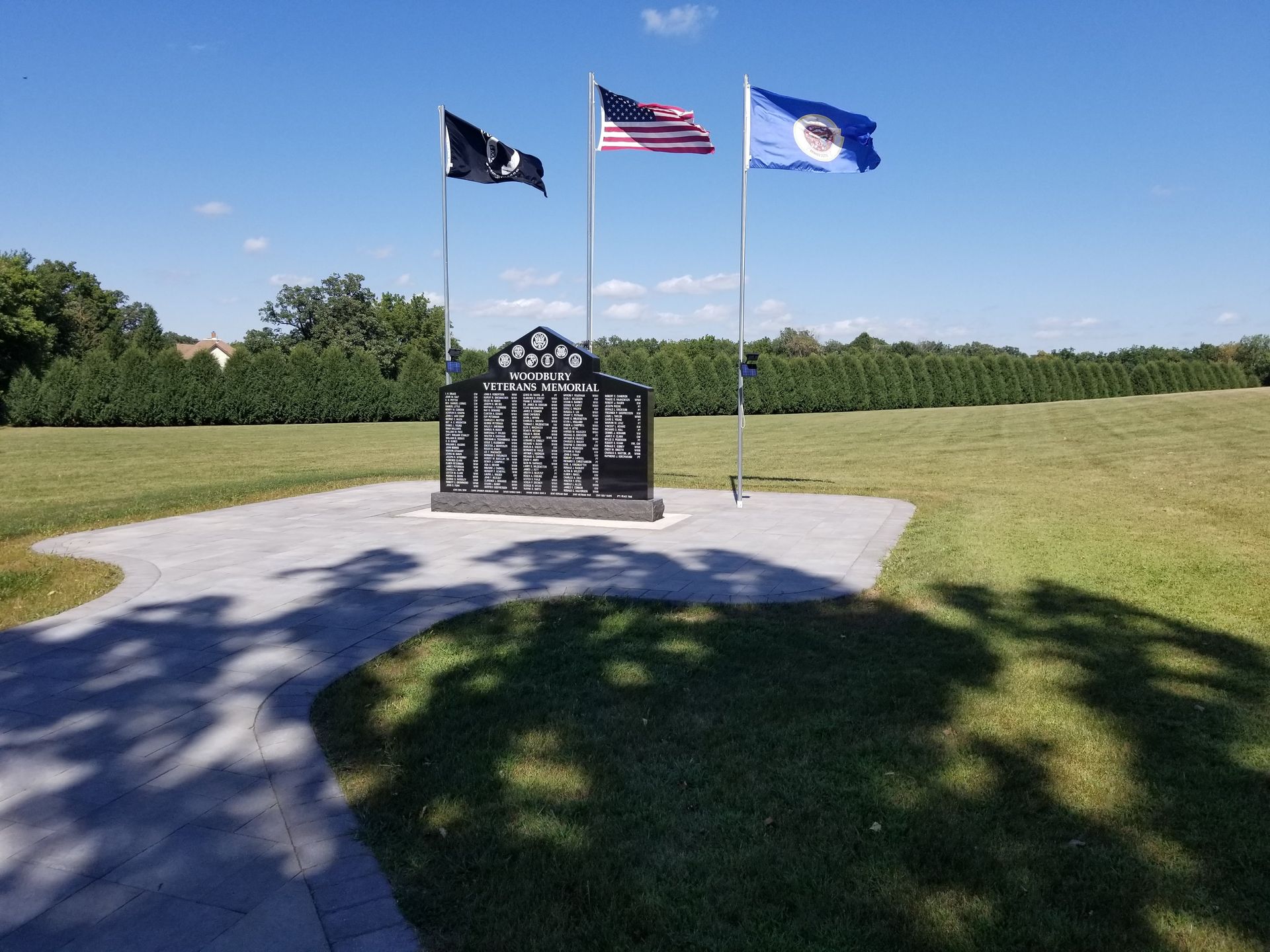 A memorial monument with lists of names, flanked by three flags on poles in a grassy field under a bright blue sky.