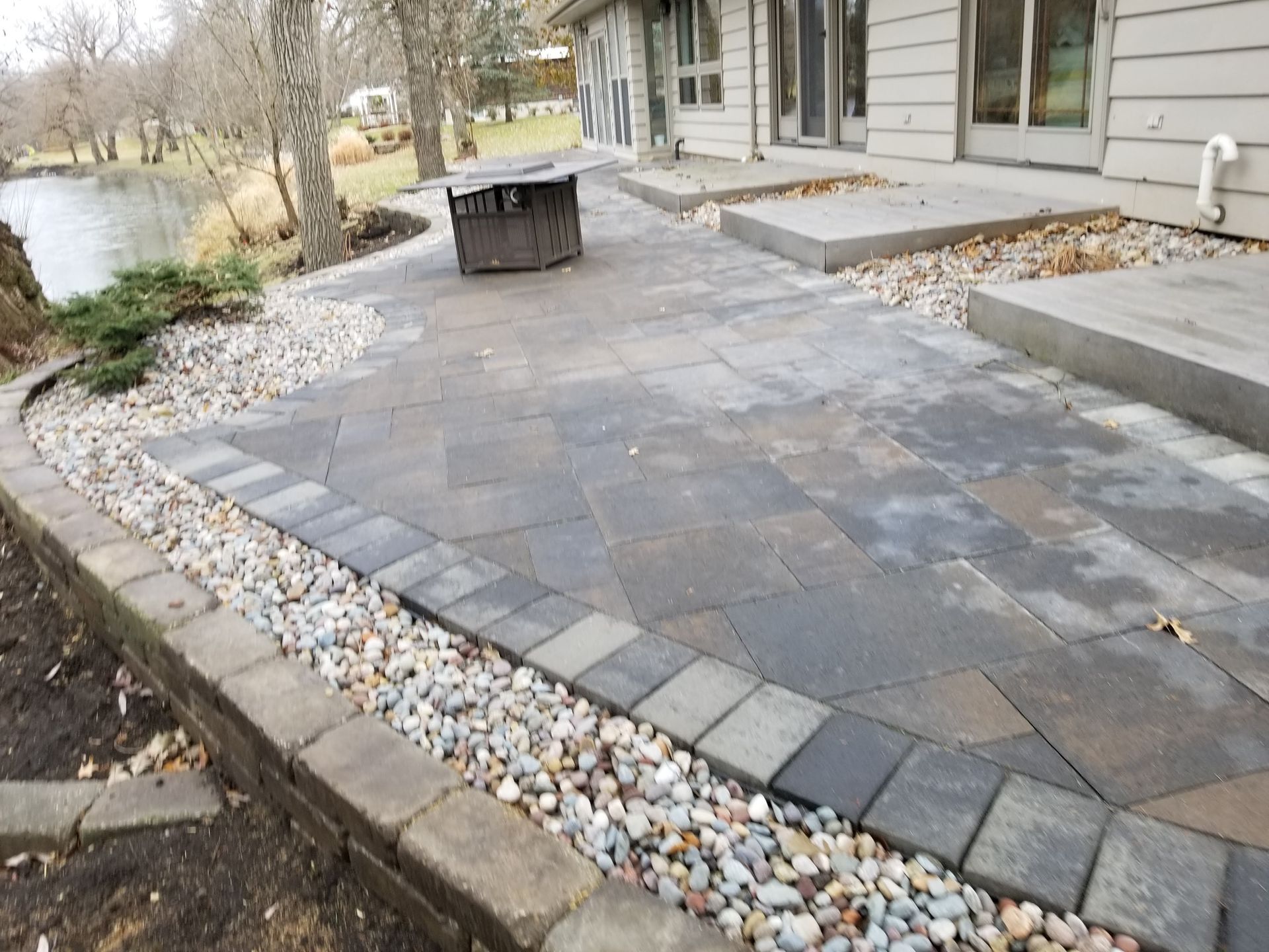 A stone patio with a fire pit beside a house and a shoreline, featuring gray pavers, a rock border, and concrete steps.
