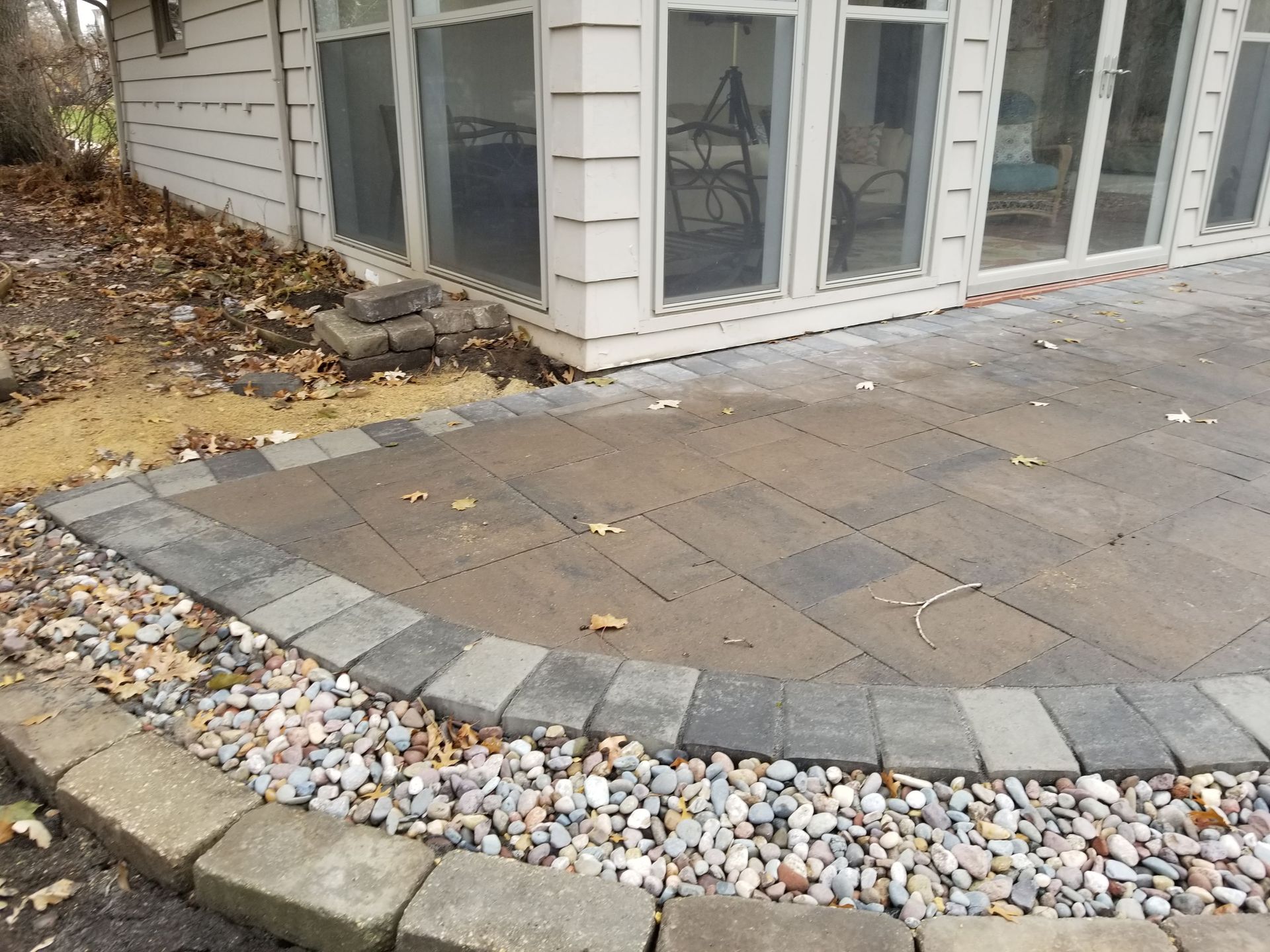 A paved stone patio with a dark grey border, surrounded by a gravel edge next to a white house with large glass windows.