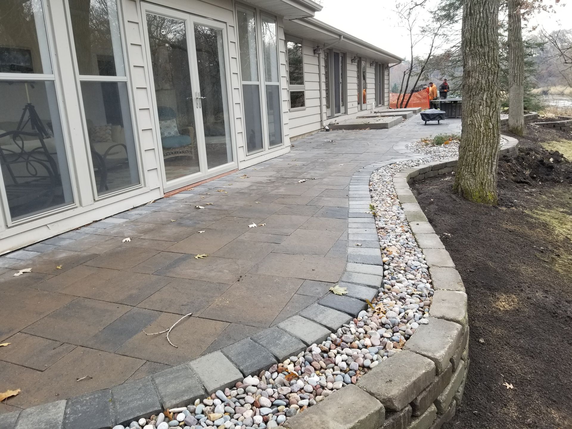 A paved patio with a curved stone retaining wall and a gravel border, located beside a house with sliding glass doors.