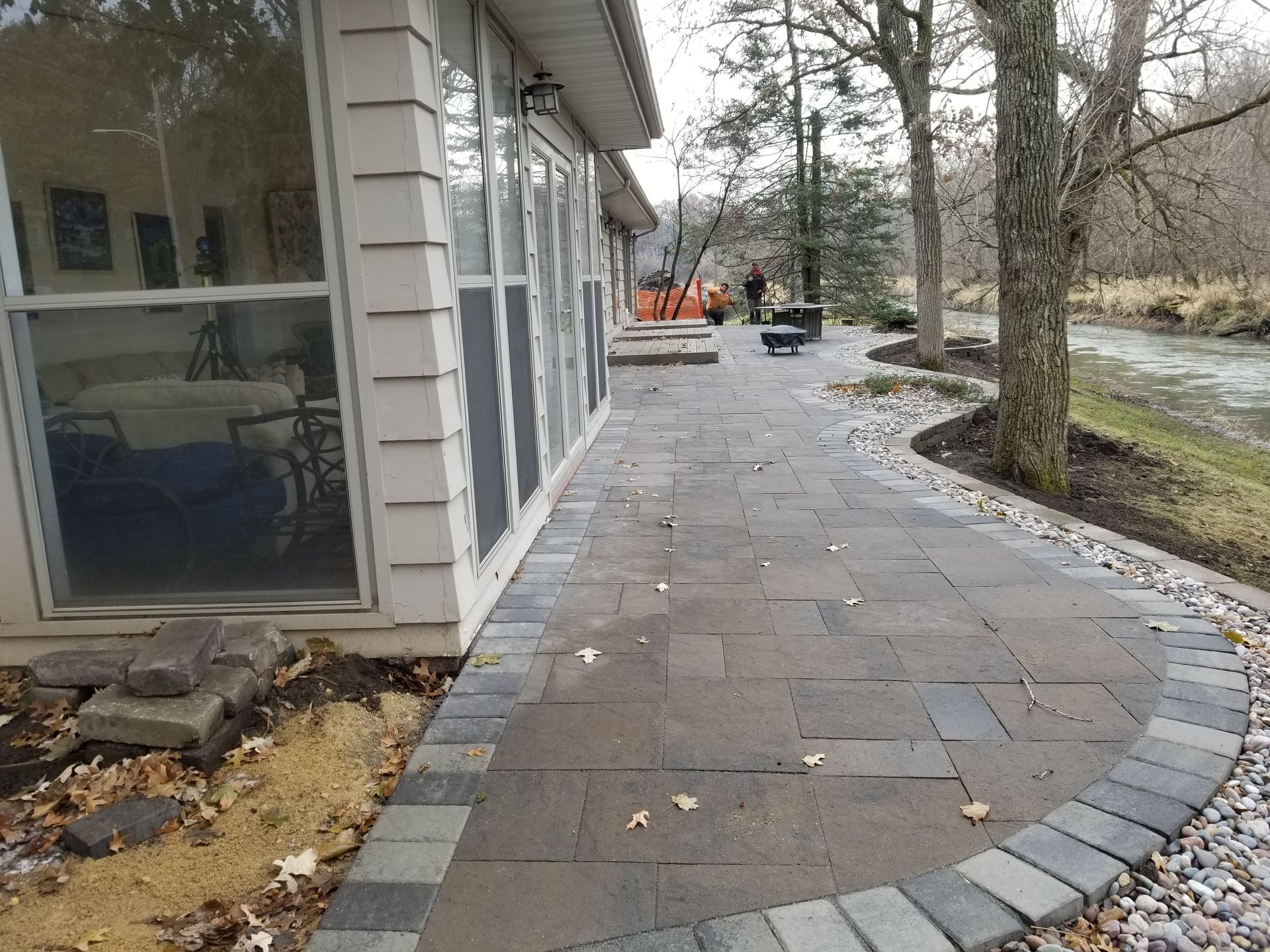 A stone patio runs alongside a house with a screened porch, leading toward a riverbank with trees in the background.