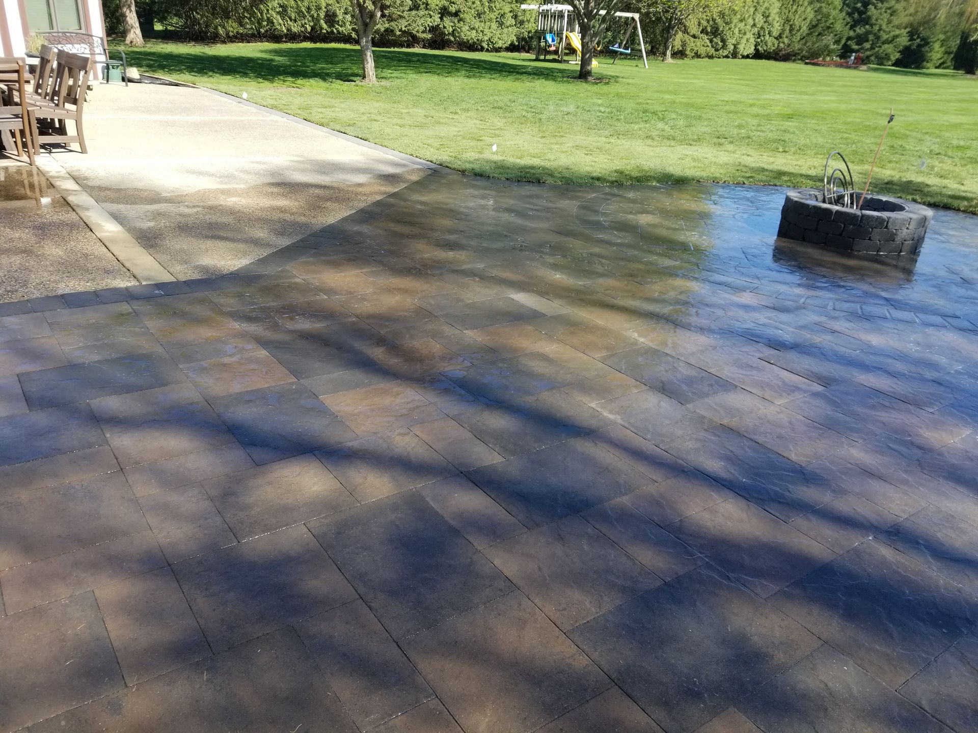 A stone patio with a fire pit ring, featuring a wet, darker section near the grass and a dry, lighter section near chairs.