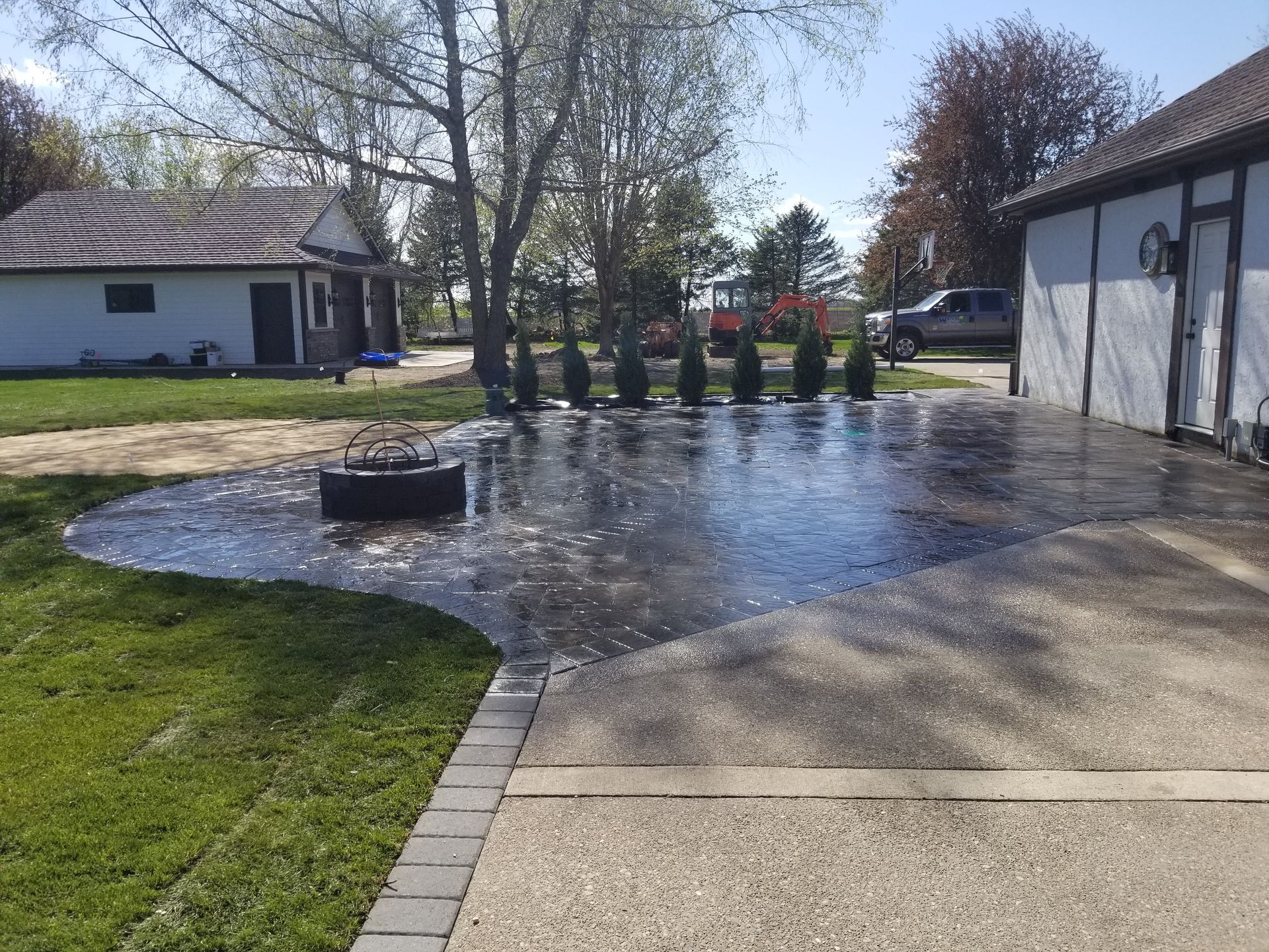 A freshly sealed, dark concrete patio with a fire pit, bordered by pavers, next to a garage on a sunny day.