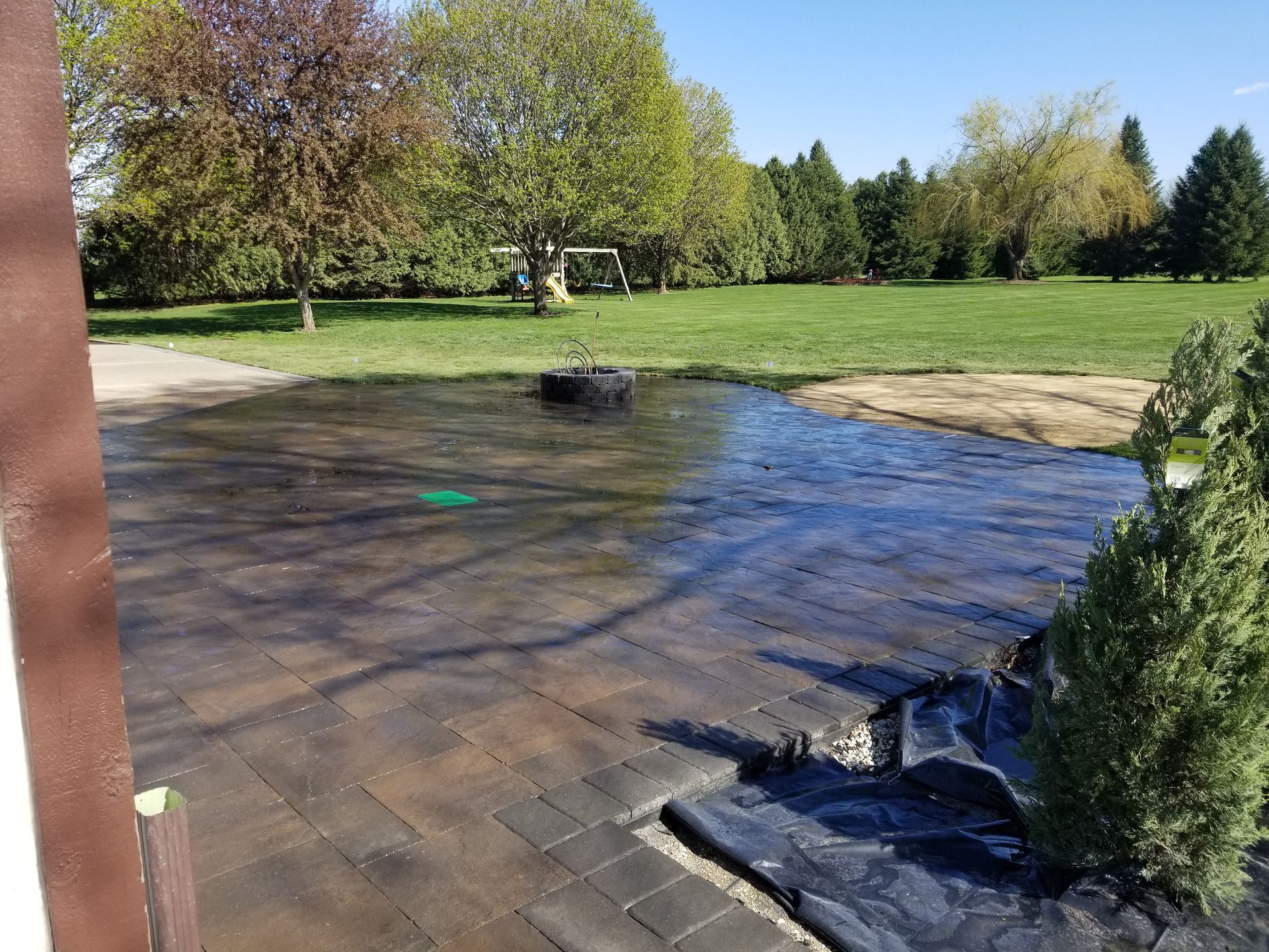 A dark stone patio under construction, with a wet surface and black landscape fabric near a green tree in a grassy yard.