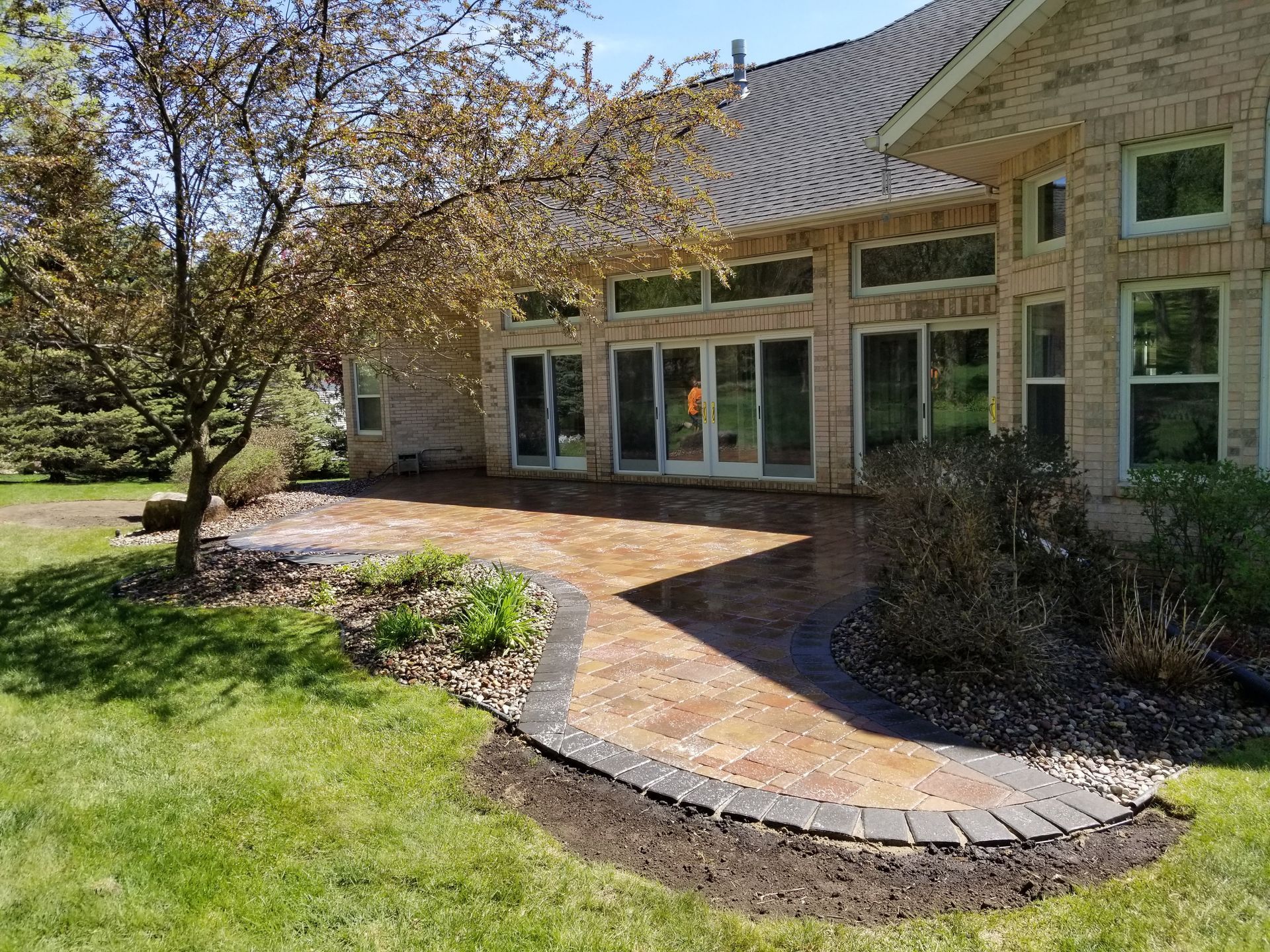 A brick patio with a dark stone border set against the back of a tan brick house with glass doors and landscaping.