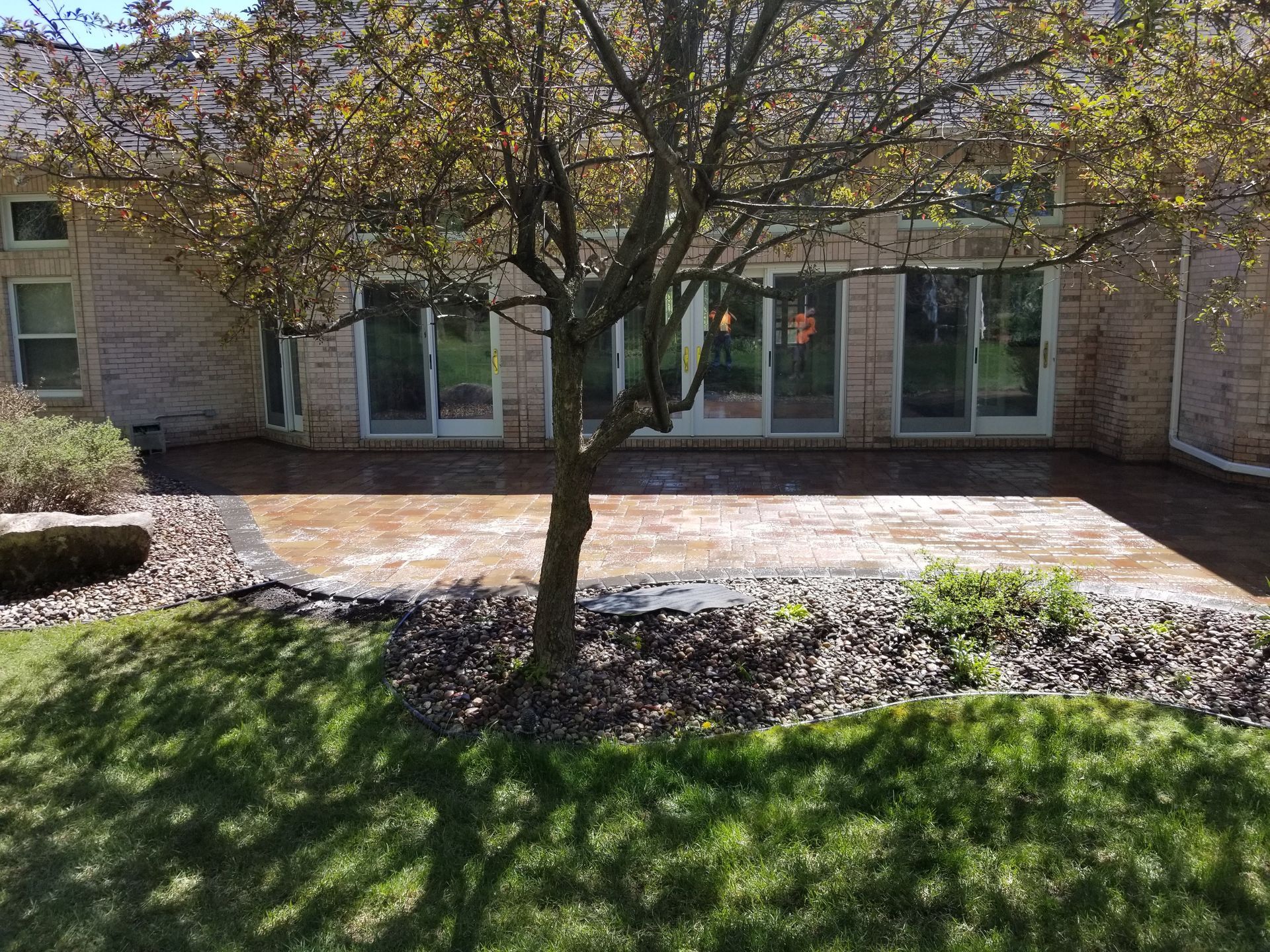 A tan brick house features a paved patio area behind a large tree, bordered by dark landscaping rocks and a green lawn.
